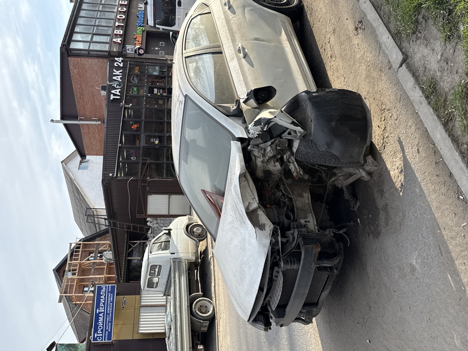 The image depicts a white, four-door sedan with extensive front-end collision damage, parked on the right shoulder of an asphalt road. The vehicle's hood is crumpled, the front bumper is dislodged, the passenger-side front fender is severely crushed and torn, and the front passenger-side wheel assembly is significantly damaged and displaced inwards. Exposed engine components and wiring are visible. A red warning triangle rests on the damaged hood.
In the background, on the opposite side of the road, a white GAZelle flatbed truck is parked. Behind the truck, there are various commercial buildings and structures. A sign for "СТРОЙМАТЕРИАЛЫ" (Building Materials) is visible on the left. To the right, another commercial building features signs including "ТАБАК 24" (Tobacco 24), "АВТОСЕРВИС" (Auto Service), and "DetaliNg StUdio L.K." (Detailing Studio L.K.). A multi-story building under construction with visible wooden scaffolding is present further back, where a single individual is visible on the scaffolding. The sky is partly cloudy, and the scene is illuminated by daylight, casting shadows towards the foreground, indicating morning or late afternoon. The location is an urban or semi-urban area in Rostov-on-Don, Russia, based on the commercial signs in Cyrillic script and the specific city requested.