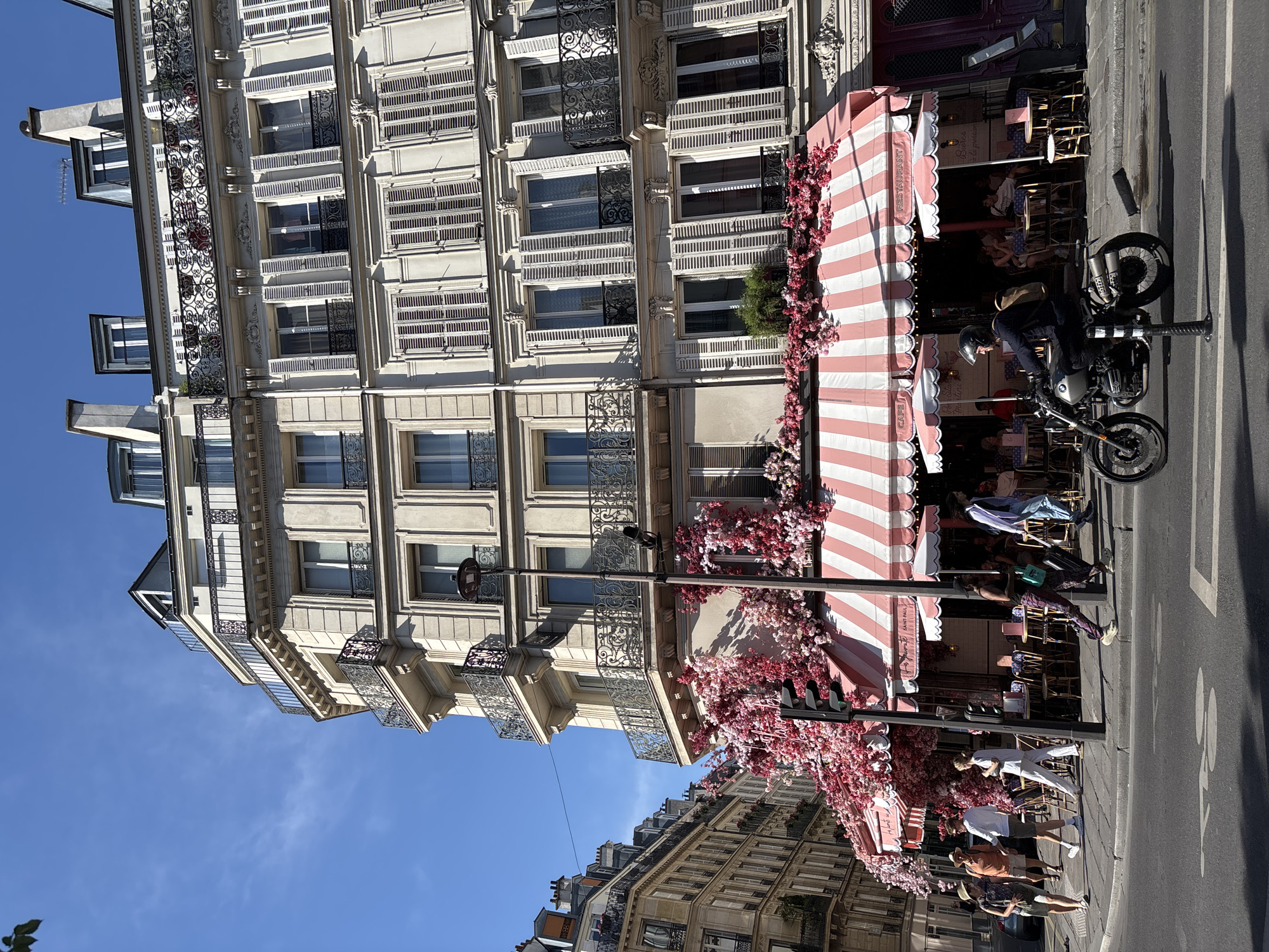 The image captures a street scene in Paris, France, on a sunny day. The focus is on a building with ornate architecture, featuring multiple stories with shuttered windows and decorative balconies. A cafe with a distinctive pink and white striped awning extends from the building, adorned with abundant pink flowers that cascade down its facade.
Several people are visible, some seated at tables outside the cafe and others walking along the sidewalk. A person on a motorcycle is parked on the street, facing the building. The overall mood is lively and urban, suggesting a pleasant day in the city.
Notable details include the clear blue sky, indicating fair weather. The architecture suggests a Parisian setting. Text is visible on the cafe awning, reading "RESTAURANT" and "SAINT-PAUL". The time of day appears to be daytime, with the sun casting shadows. The scene evokes a sense of everyday life in a vibrant European city.