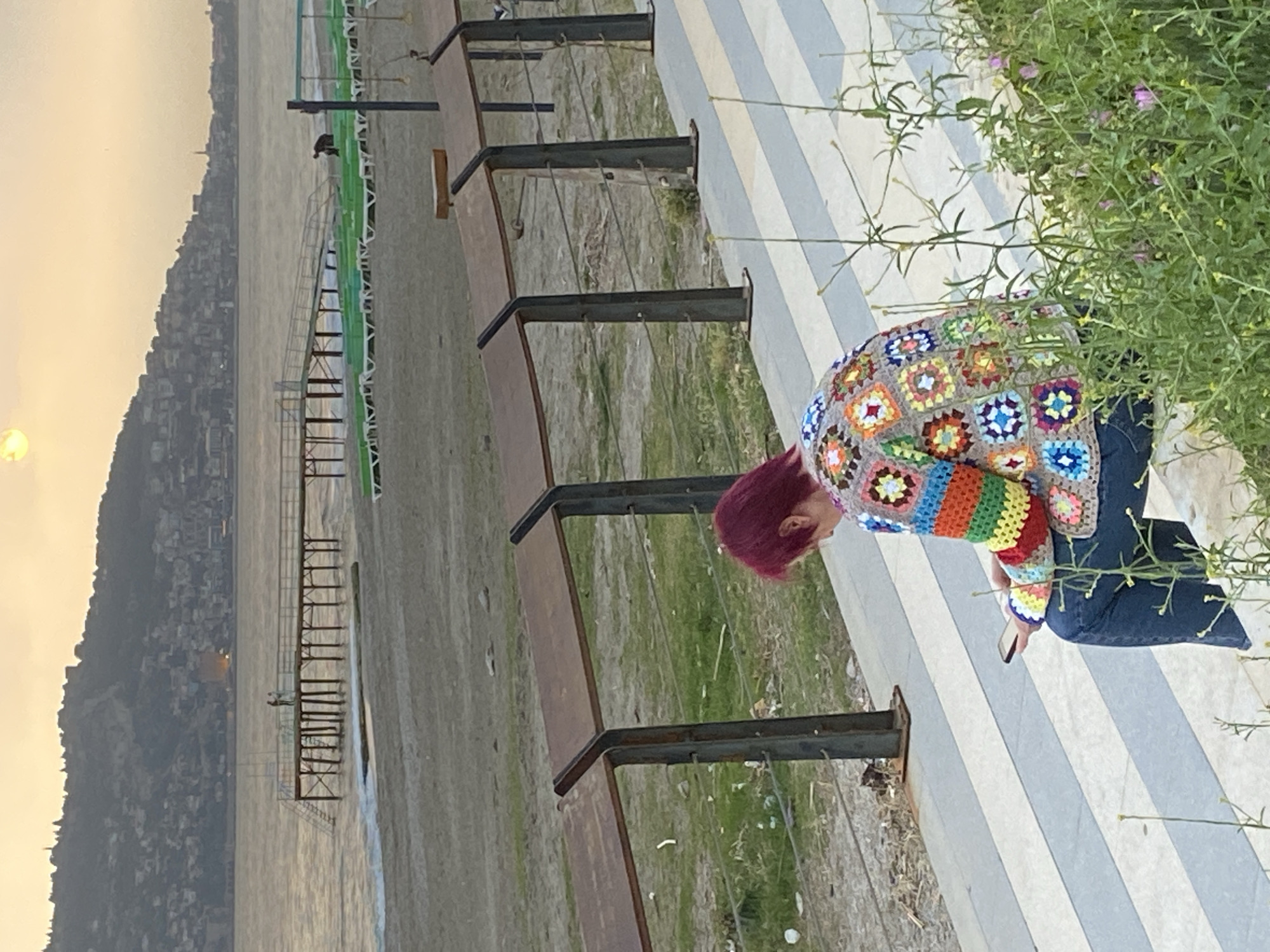A medium shot depicts a red-haired person seated on a bench, facing forward and slightly downwards at their mobile phone. The individual is dressed in blue jeans and a colorful crochet cardigan composed of rainbow-colored granny squares. The person is positioned on a bench with metal supports and brown wooden seating, next to a sidewalk featuring alternating wide stripes of white and light gray. Green plants with small purple and yellow flowers are visible at the edge of the sidewalk. The background shows a beach, a metal pier-like structure leading into the water, and across the water, a hillside covered with buildings under a hazy sky. This setting is consistent with the beach of Alanya, Turkey, given the topography and structural features.