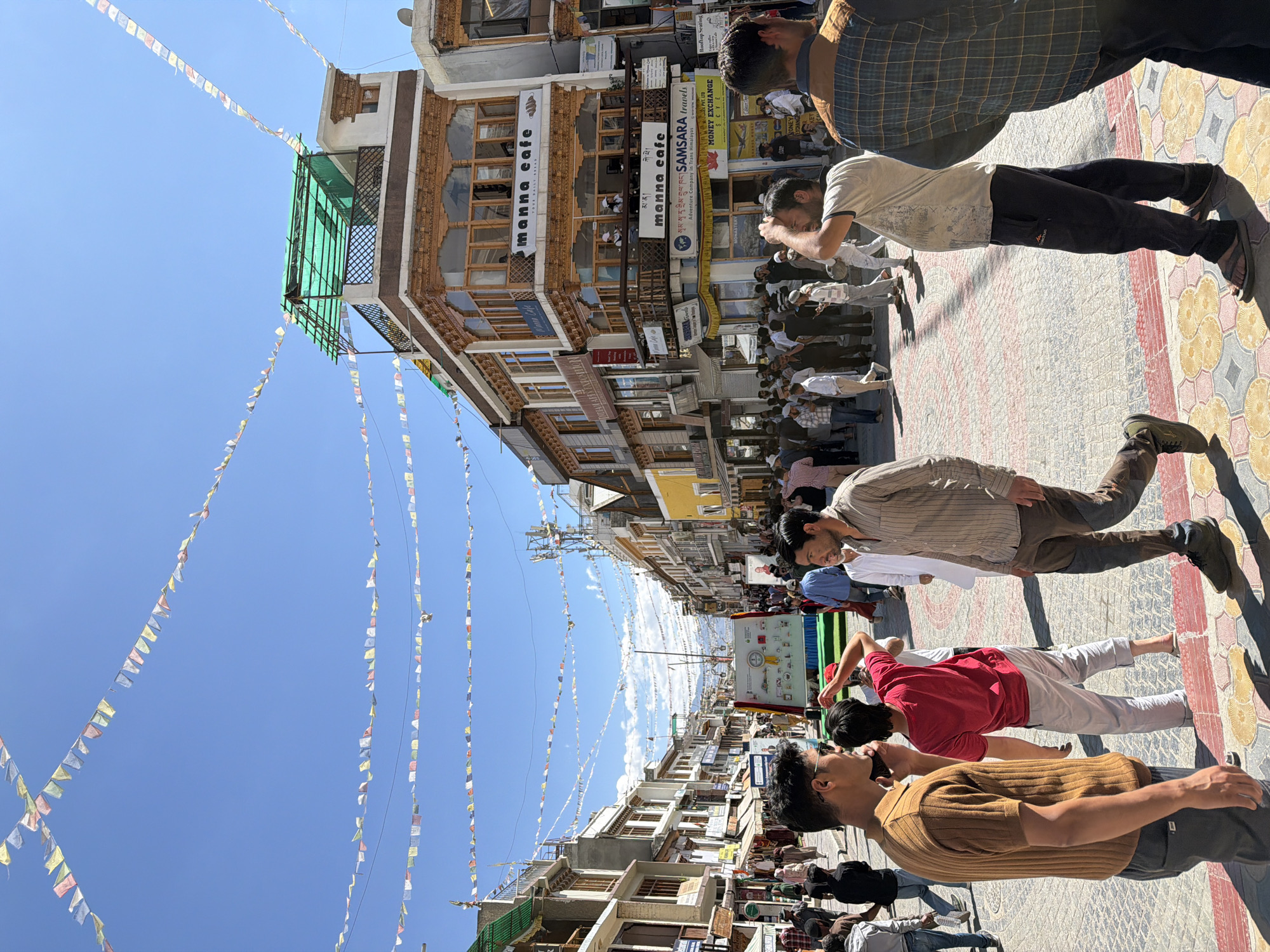 This is a medium-shot, eye-level, outdoor photograph taken on a sunny day in Leh, India. The scene depicts a bustling street with a crowd of people walking and gathered. Prayer flags are strung across the sky, creating a festive atmosphere. Buildings line both sides of the street, featuring traditional Tibetan architecture with wooden balconies and intricate carvings. The buildings house various businesses, including shops, cafes, and travel agencies.
In the foreground, several individuals are visible, including a man in a brown ribbed t-shirt and dark pants, another man in a red t-shirt and white pants, and a man in a striped shirt and brown pants. In the background, a large banner with various symbols and text is displayed, likely related to an event or information. People are seen in different attire, suggesting a mix of locals and tourists. The street is paved with colorful tiles. The overall impression is that of a lively and vibrant street scene in the heart of Leh.