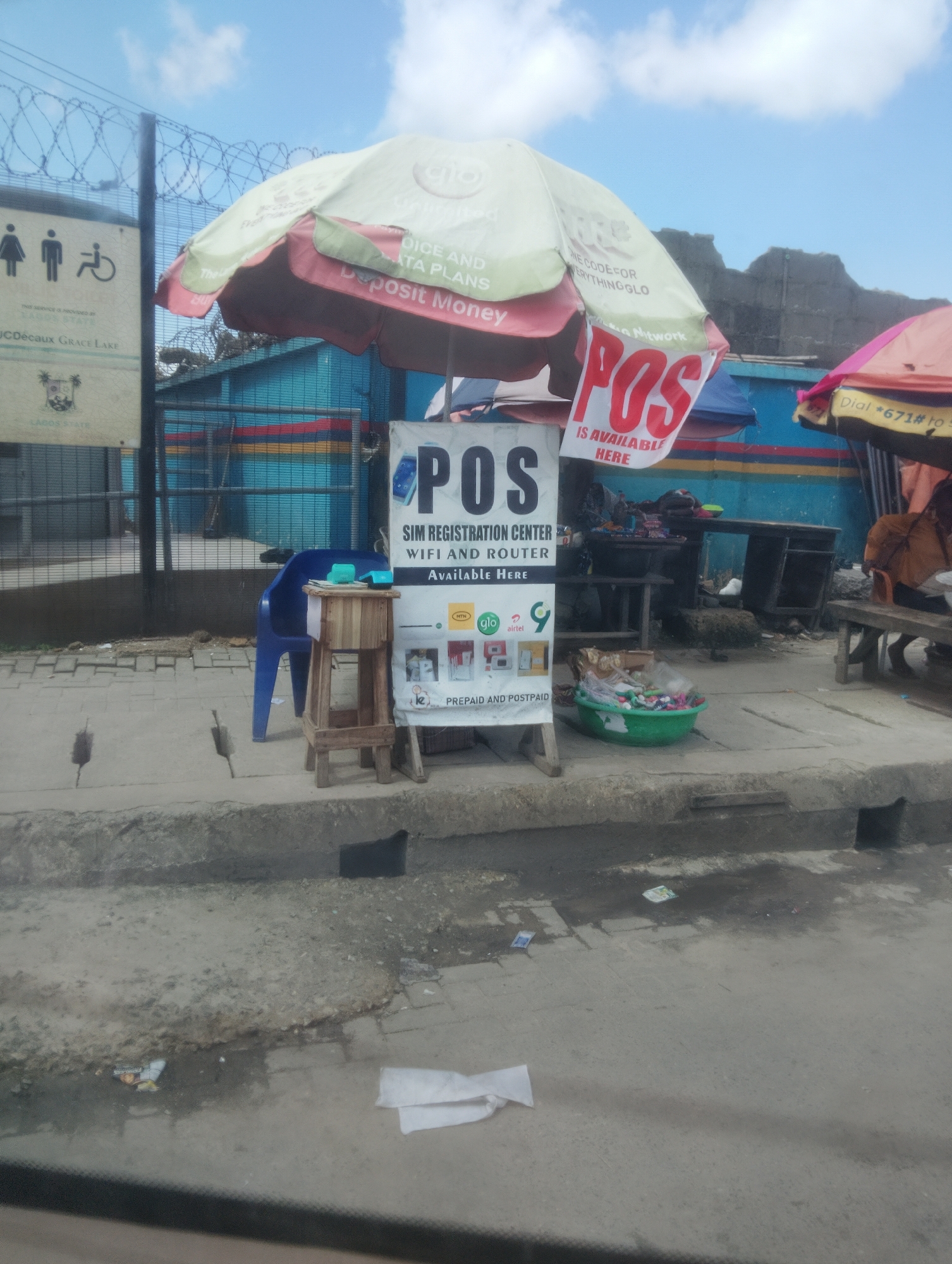 The image depicts a street scene in Oworonshoki, Nigeria, under a cloudy sky. In the foreground, a concrete sidewalk and gutter are visible, with some debris scattered on the ground.
The main subject is a roadside stall or booth. A large, open umbrella, predominantly white and red, is set up to provide shade. A sign hanging from the umbrella reads "POS IS AVAILABLE HERE" in large red letters. Below this, another sign indicates "SIM REGISTRATION CENTER, WIFI AND ROUTER AVAILABLE HERE". This sign also features logos for mobile network providers like GLO and Airtel, along with images related to prepaid and postpaid services.
Adjacent to the POS stall, a simple wooden stand supports a small object, possibly a cash register or payment terminal. A blue plastic chair is placed nearby.
In the background, a chain-link fence with barbed wire runs across the scene. Behind the fence, a blue building with colorful horizontal stripes is partially visible. Further back, the landscape appears somewhat rugged with hints of other structures. Another umbrella, partially visible on the right side of the image, is a vibrant pink, suggesting another vendor or stall.
The scene suggests a typical urban commercial environment, likely a busy area where people can access mobile services and make electronic transactions. There are no people clearly visible in the frame.