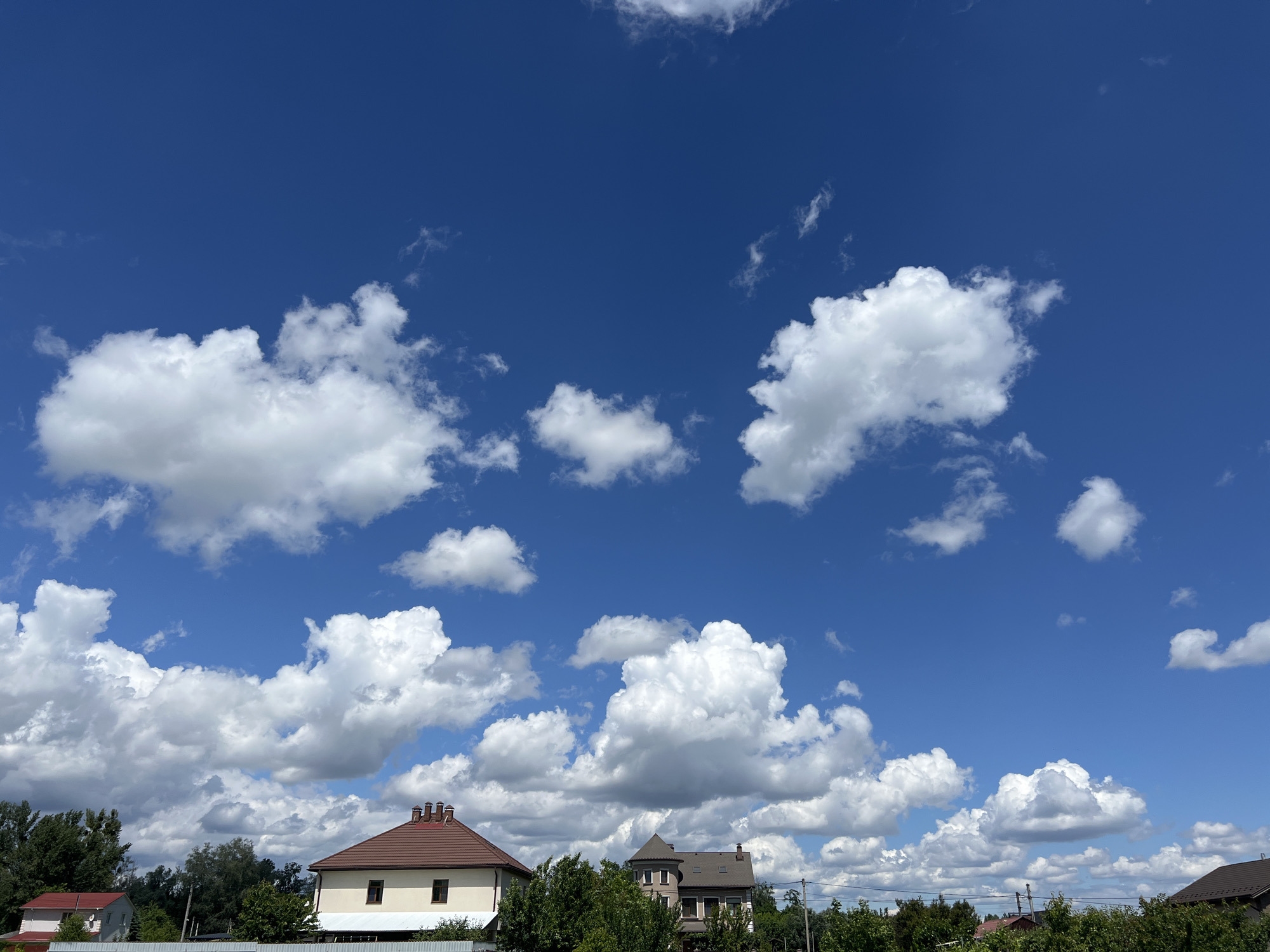The image shows a bright blue sky with fluffy white cumulus clouds scattered across it. Below the sky, there are several residential buildings, trees, and a fence. On the right side of the image, a house with a brown tiled roof and cream-colored walls is visible, featuring several chimneys. To its left, another smaller house with a red roof can be seen. The scene suggests a suburban or rural residential area. This image was taken in the city of Boyarka, Ukraine.