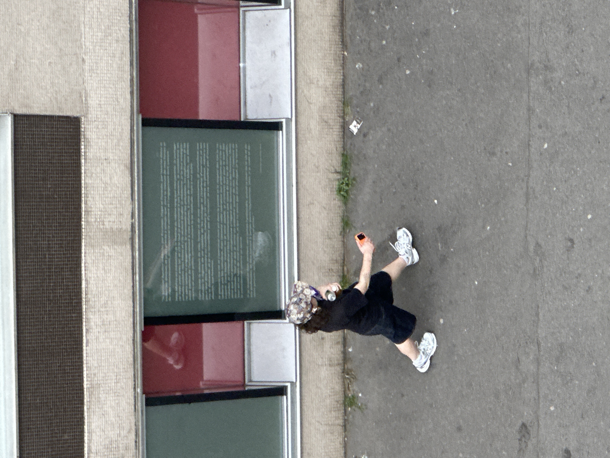 An aerial, high-angle view captures a person walking alongside a building facade. The person, likely female, is seen from above, moving from the bottom-right towards the upper-left of the frame. She wears a patterned cap, a dark t-shirt, dark shorts, and white sneakers, holding an orange-cased smartphone in her right hand. A white earbud or wire is visible near her left ear.
The building on the left features a multi-paneled exterior. Visible elements include sections of light-colored, textured material (possibly stone or concrete), dark mesh-like panels, and rectangular red panels. Several large, green-tinted windows are also prominent, with one large window displaying a substantial block of white text, suggesting an informational display or artwork, though the text itself is illegible due to the angle and distance. Reflected shapes, possibly of hands or other objects, are faintly visible in some of the red panels.
The ground on the right side of the image is a grey asphalt or concrete surface, showing minor wear and some scattered small debris. Small patches of green vegetation grow along the edge where the pavement meets the building. The lighting suggests an overcast daytime, as there are no harsh shadows. The location context is Paris, France.