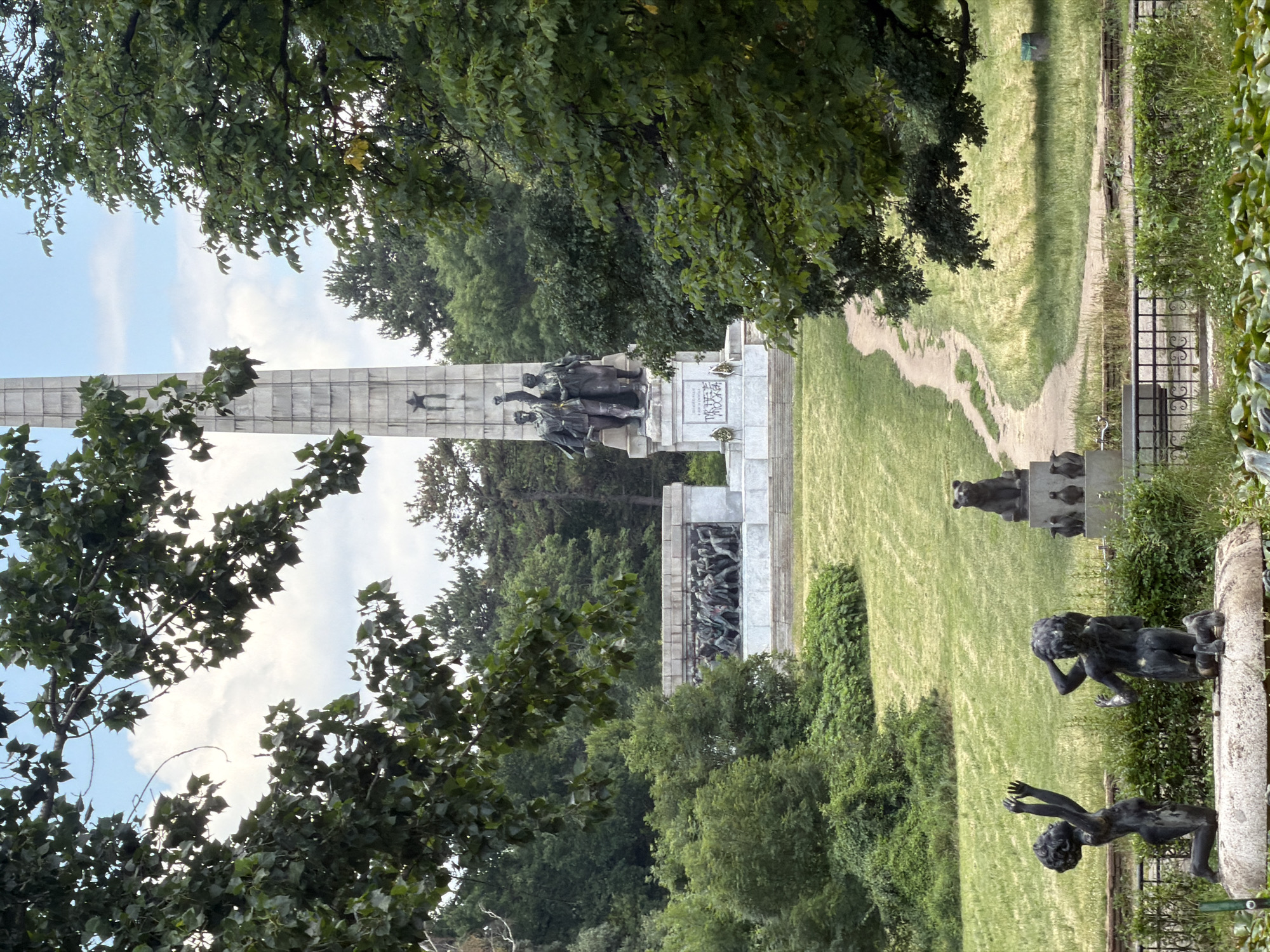 This is a full, eye-level, outdoor, daytime photograph of the Monument of the Soviet Army in Sofia, Bulgaria. The monument is situated on a hill and is partially obscured by trees in the foreground. In the midground, a large obelisk with figures of Soviet soldiers stands atop a tiered pedestal. To the left of the obelisk, a bas-relief depicts a scene of soldiers in battle, and to the right, more trees and foliage are visible. In the foreground, on the left, two dark bronze sculptures of children are depicted, one kneeling with arms raised, and the other kneeling with hands over its head. Behind them, atop a pedestal, is a bronze sculpture of a bear and several penguins. A path winds up the grassy slope toward the monument. The sky is partly cloudy, with patches of blue visible through the leaves of the trees. The overall impression is of a solemn historical site set within a green, park-like environment.