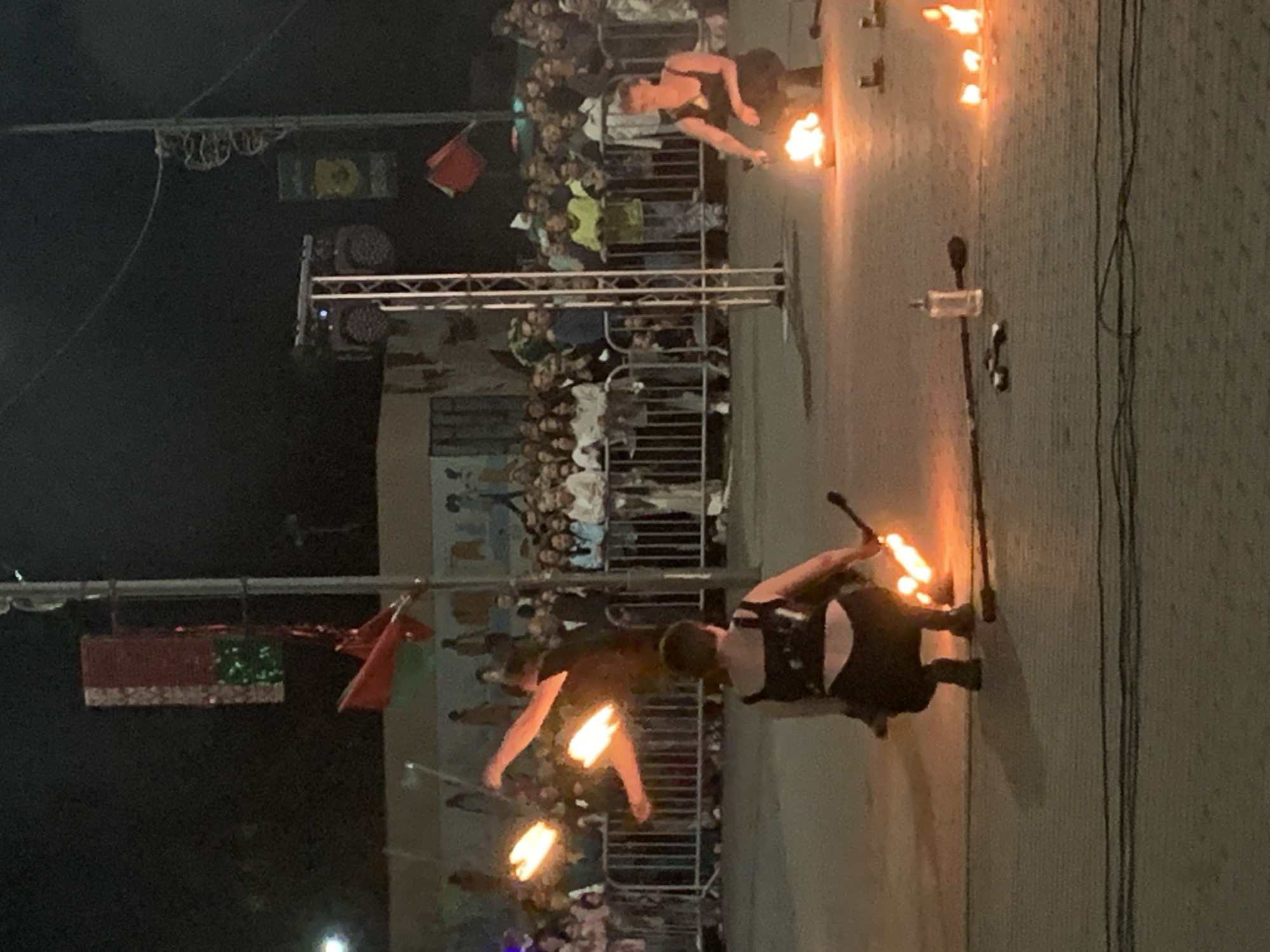 The image depicts an outdoor nighttime performance involving fire spinners in the city of Kapyĺ, Belarus. In the foreground, two individuals are actively engaged in fire spinning. The person on the left, viewed from behind, wears a black outfit and is spinning what appears to be a fire staff. Flames are visible and emanating from the staff. The person on the right, facing the camera, is crouched and also appears to be spinning a fire prop. Several burning fire elements are scattered on the ground in front of them.
In the background, a crowd of spectators is gathered behind a metal barrier, observing the performance. The environment appears to be a public space, possibly a street or plaza, with various elements suggestive of a festival or event. Visible structures include a tall metal pole with what looks like a banner or decoration, and a truss structure with lights. In the far background, buildings and illuminated elements are faintly visible against the dark night sky. The overall scene suggests a lively and dramatic entertainment event taking place in Kapyĺ.