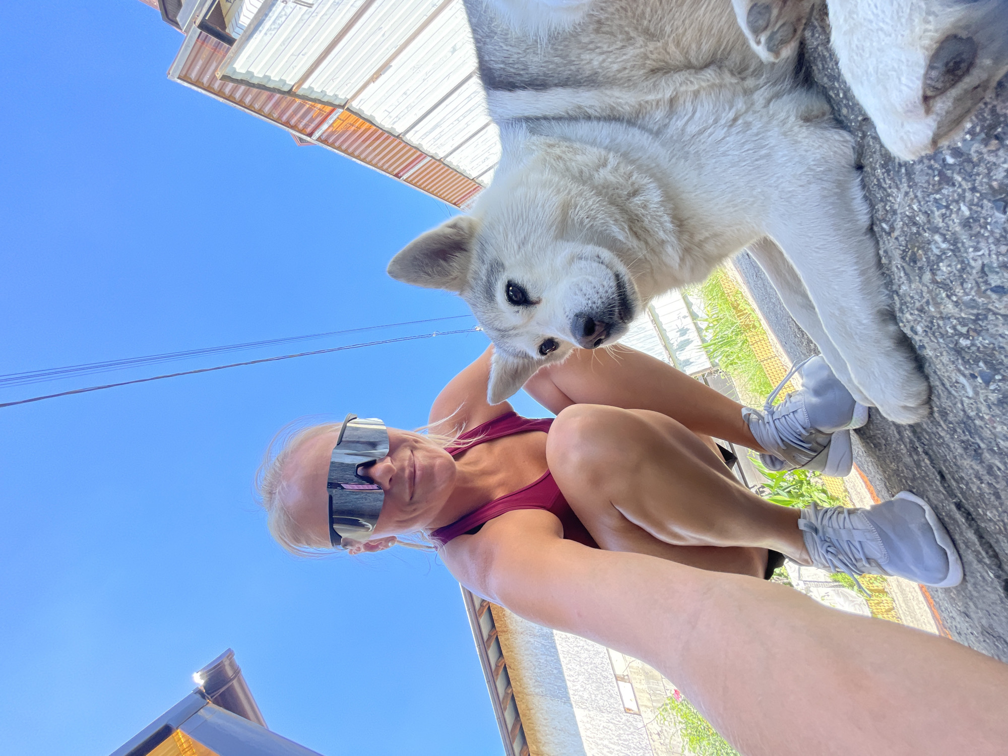 A woman wearing large, dark sunglasses and a maroon tank top squats down outdoors, facing the camera. Next to her, a light-colored dog with grey markings lies on the ground. The woman's left arm extends towards the camera, as if taking a selfie. Her legs are bent, and she wears grey athletic shoes. The background shows a clear blue sky, some overhead wires, and buildings with corrugated metal roofs. The ground appears to be concrete or asphalt. The location is identified as Baranovka, Russia.