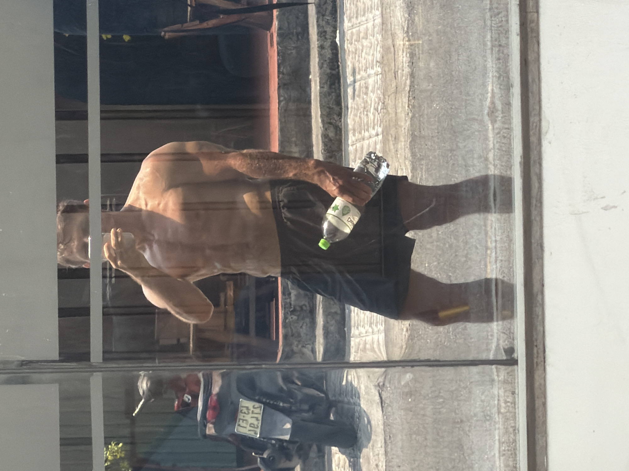 The image shows a reflection in a window. A shirtless man is holding a water bottle. The reflection also shows a motorcycle parked outside with a license plate that reads "SJRAR 13-E1", which indicates a location in the city of Đà Nẵng, Vietnam.