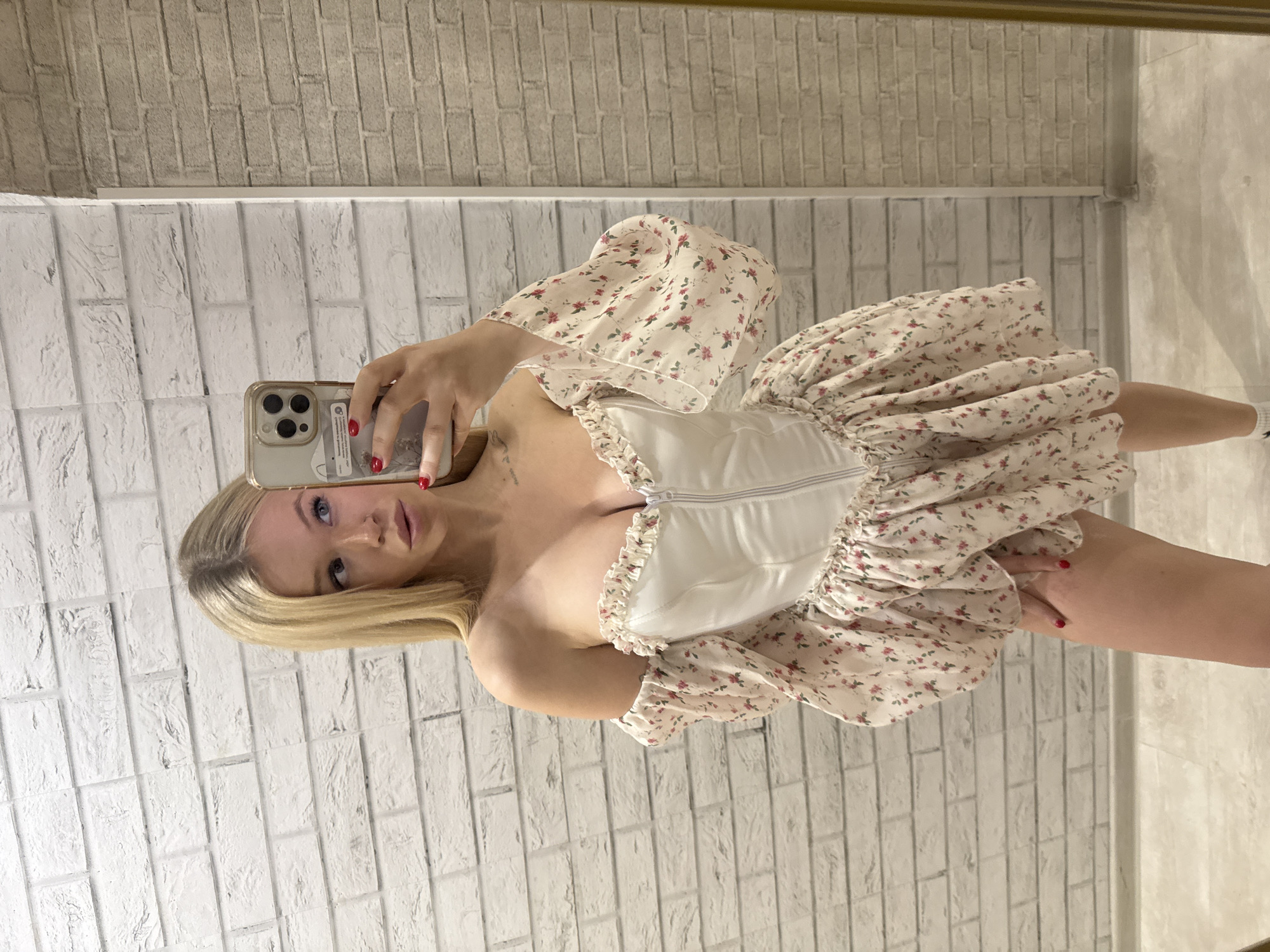 A young woman with blonde hair is taking a mirror selfie in what appears to be a home setting. She is wearing a white corset top with a floral print skirt that has puff sleeves. The background consists of a white brick wall. She is holding a smartphone in her left hand, which is capturing the reflection. Her facial expression is neutral. The lighting suggests it is daytime, possibly indoors with artificial lighting. There are no clear indications of weather or specific time of day beyond that. The location context provided is Saint Petersburg, Russia, but there are no visible cues in the image to confirm this.