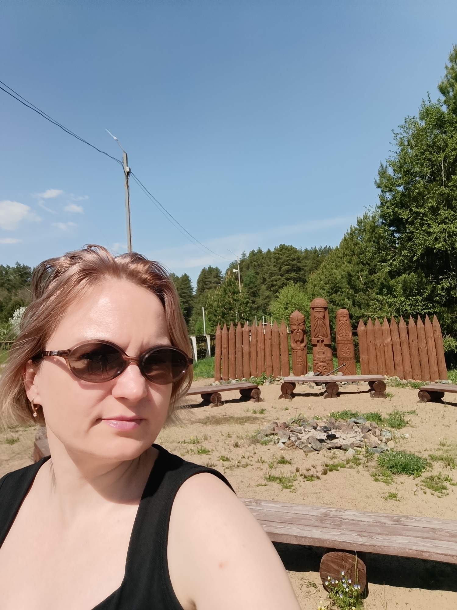 The photograph depicts a woman wearing sunglasses and a black tank top, likely taking a selfie. She is positioned in front of a scene featuring a wooden fence, three carved wooden figures, and benches arranged around a fire pit. The ground is sandy. In the background, there is a forest and a clear blue sky with a few clouds. A utility pole with wires is visible in the upper left corner of the frame. The location is identified as Prikamskii, Russia, based on contextual information.