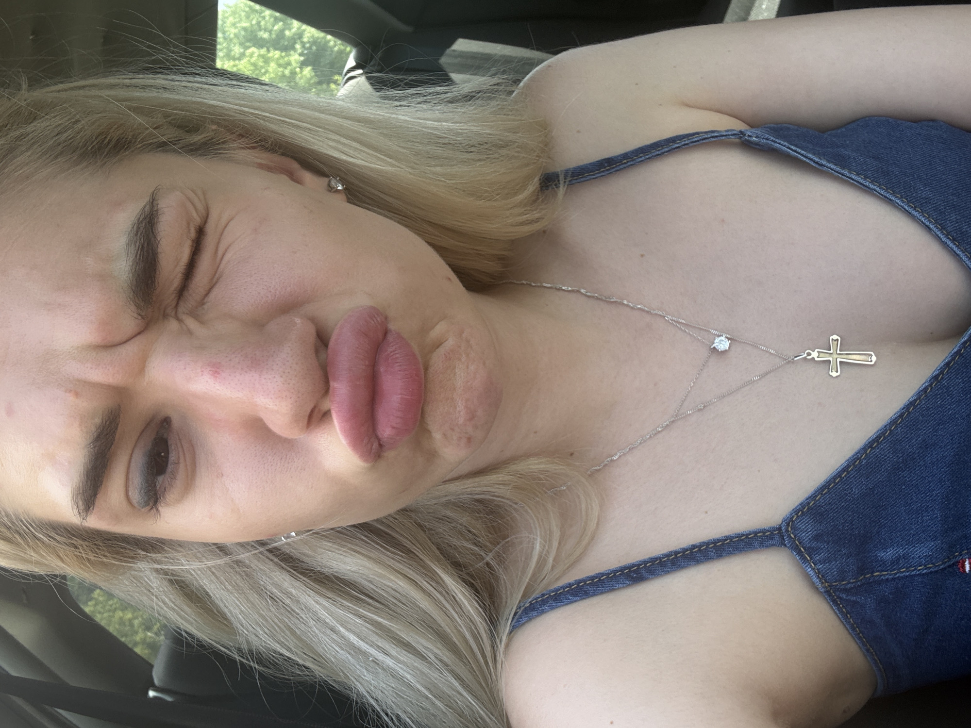 A young blonde woman is depicted in a close-up, chest-up portrait, making an exaggerated facial expression. She has light skin with visible pores and some minor redness or blemishes on her nose and chin. Her lips are pursed forward in a "duck face," and her right eye (viewer's left) is completely closed in a wink, while her left eye (viewer's right) is squinted, causing wrinkles around the eyes and forehead. She wears a blue denim-style top with thin straps and two silver-toned necklaces; one with a small clear gemstone pendant and another with a gold-colored cross pendant. A silver stud earring is visible in her right ear (viewer's left). The background suggests she is inside a vehicle, possibly a car, with dark interior elements visible behind her. Through a window, green foliage (trees) is visible under bright, natural lighting. The image does not contain any visual information to specify the location as Novo Konjarevo, North Macedonia.