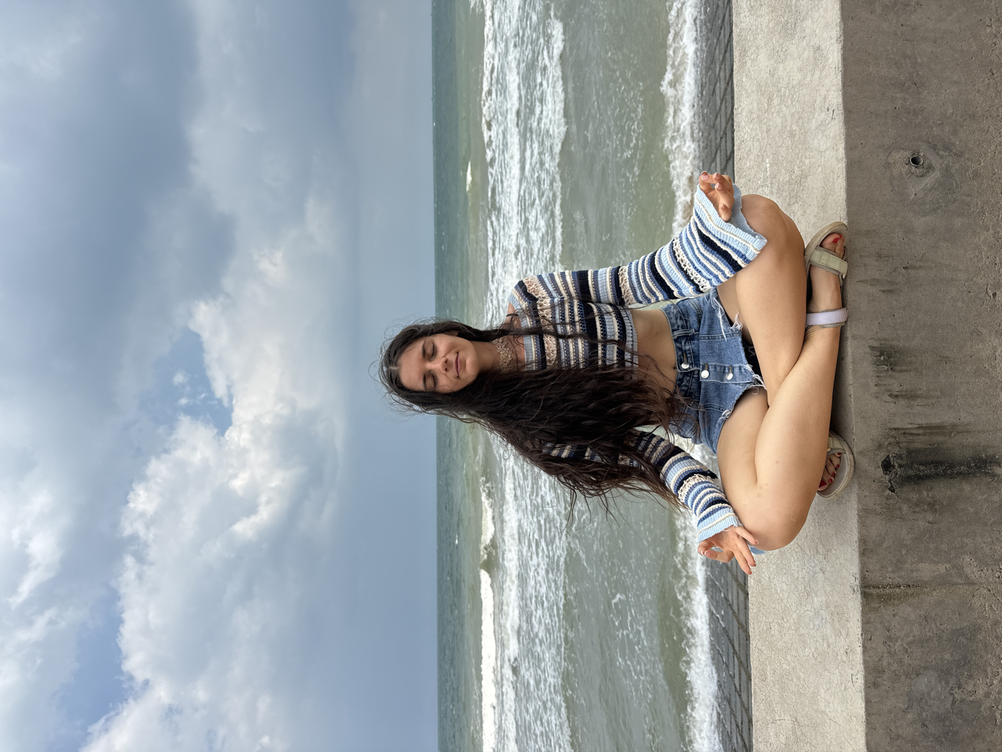 A young woman with long, dark hair is meditating in a lotus position on a concrete barrier overlooking the sea. She is wearing a striped, cropped sweater and denim shorts. The ocean waves are visible behind her, and the sky is overcast. The scene appears to be in Phu Thuy Ward, Vietnam.