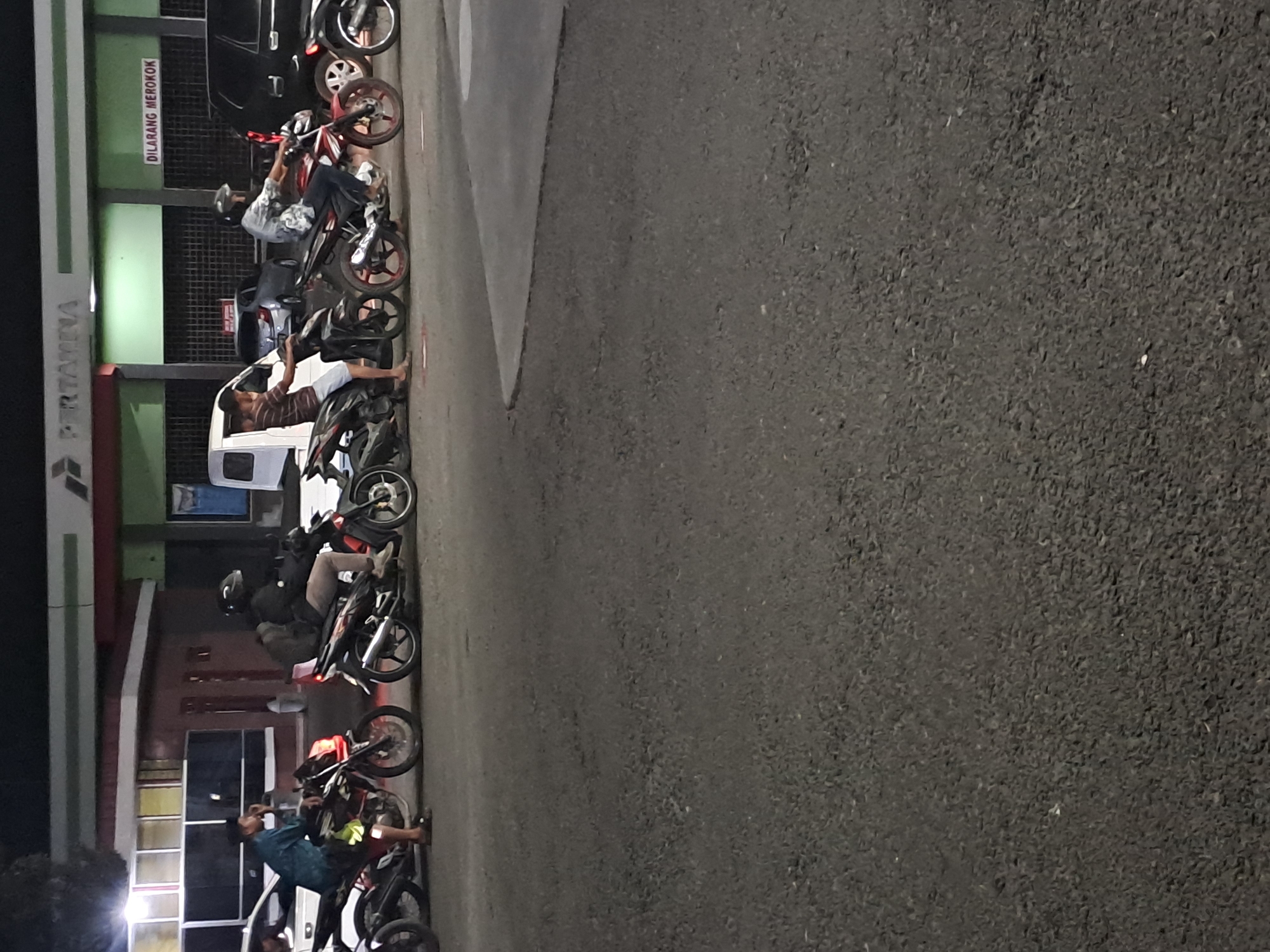 At dusk, several motorcycles are parked on asphalt in front of a Pertamina gas station in Slambur, Indonesia. Two individuals are seated on motorcycles, one of whom is wearing a helmet and a patterned shirt. Another person, standing and wearing a blue shirt, is leaning on a motorcycle. The gas station building has a green facade and a red trim. A white van and a black car are also visible. A sign on the gas station reads "DILARANG MEROKOK" which translates to "NO SMOKING". The ground is a dark asphalt surface.