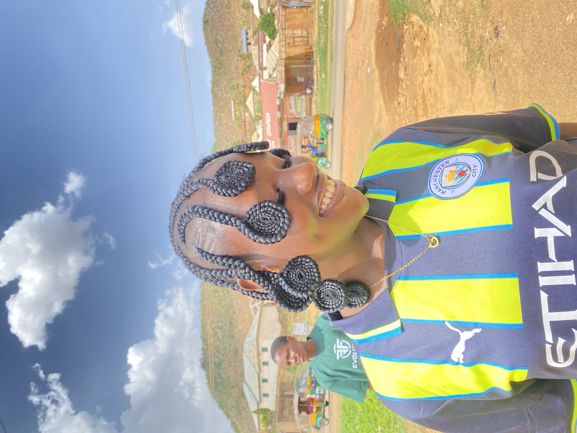 A medium shot depicts a young woman with intricately braided hair smiling brightly. Her hair is styled with braids that coil into circular shapes near her forehead and temples. She wears a navy blue Manchester City soccer jersey with bright yellow and light blue stripes and a "ETIHAD" sponsor logo on the front. She also has a gold chain necklace with a heart pendant. The image appears to be rotated to the left.
In the background, there is a hilly landscape with buildings and vegetation. A person wearing a green T-shirt can be seen slightly blurred in the midground. Further back, an "CHURCH" sign is seen above a road in Gwong, Nigeria.