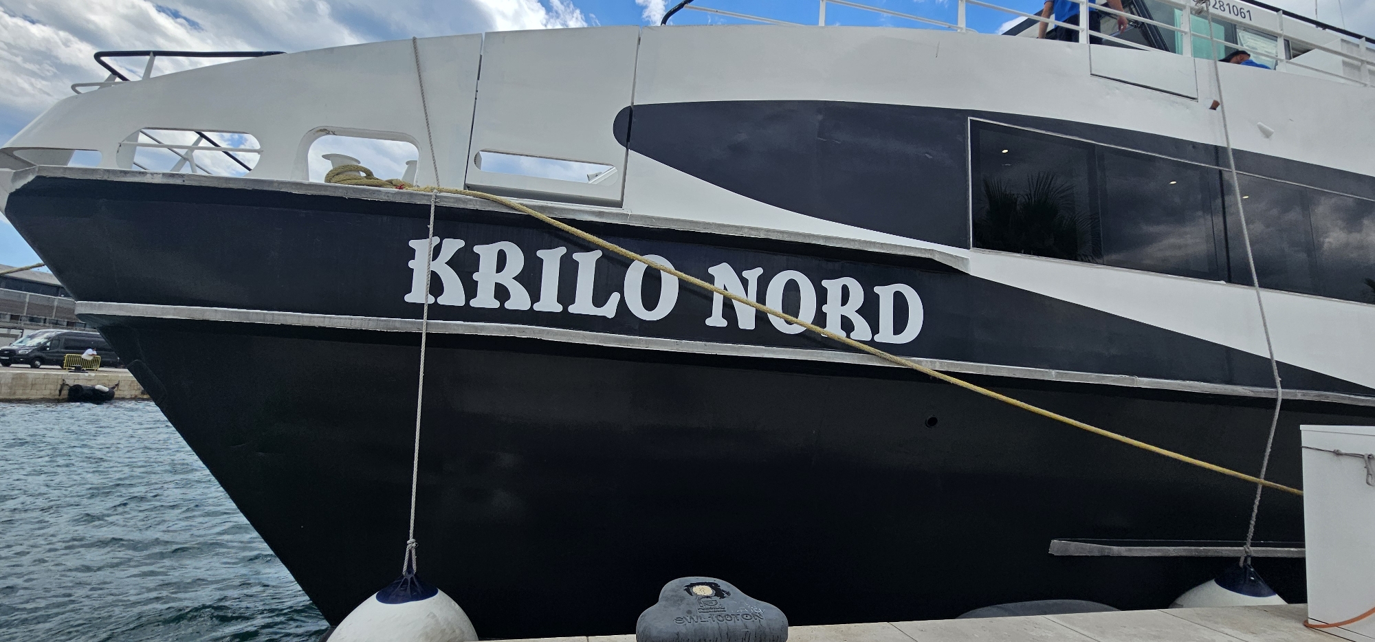 The image shows a close-up of the side of a large black and white boat named "KRILO NORD" docked in the city of Split, Croatia. The boat has a black hull with white lettering and a white upper structure. The name "KRILO NORD" is prominently displayed in large, white, stylized letters on the black hull. The boat is tethered to a dock with thick ropes and has fenders hanging over the side. In the background, there are reflections of palm trees in the windows of the boat and a cloudy sky above. To the left, a dark-colored van and a yellow barrier are visible near the dock. The water surrounding the boat is blue and appears calm.