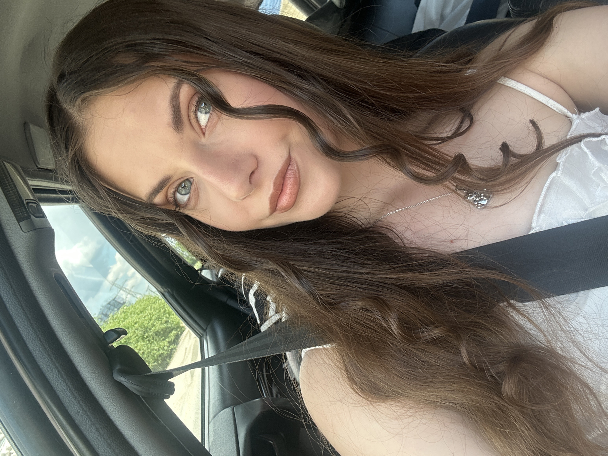 A young woman with long brown hair, fair skin, and blue eyes sits in a car. She wears a white top with thin straps and a silver necklace. A black seatbelt is fastened across her chest. The car's interior is black. Through the window, greenery and buildings are visible in the distance. The city is Unknown, Unknown.