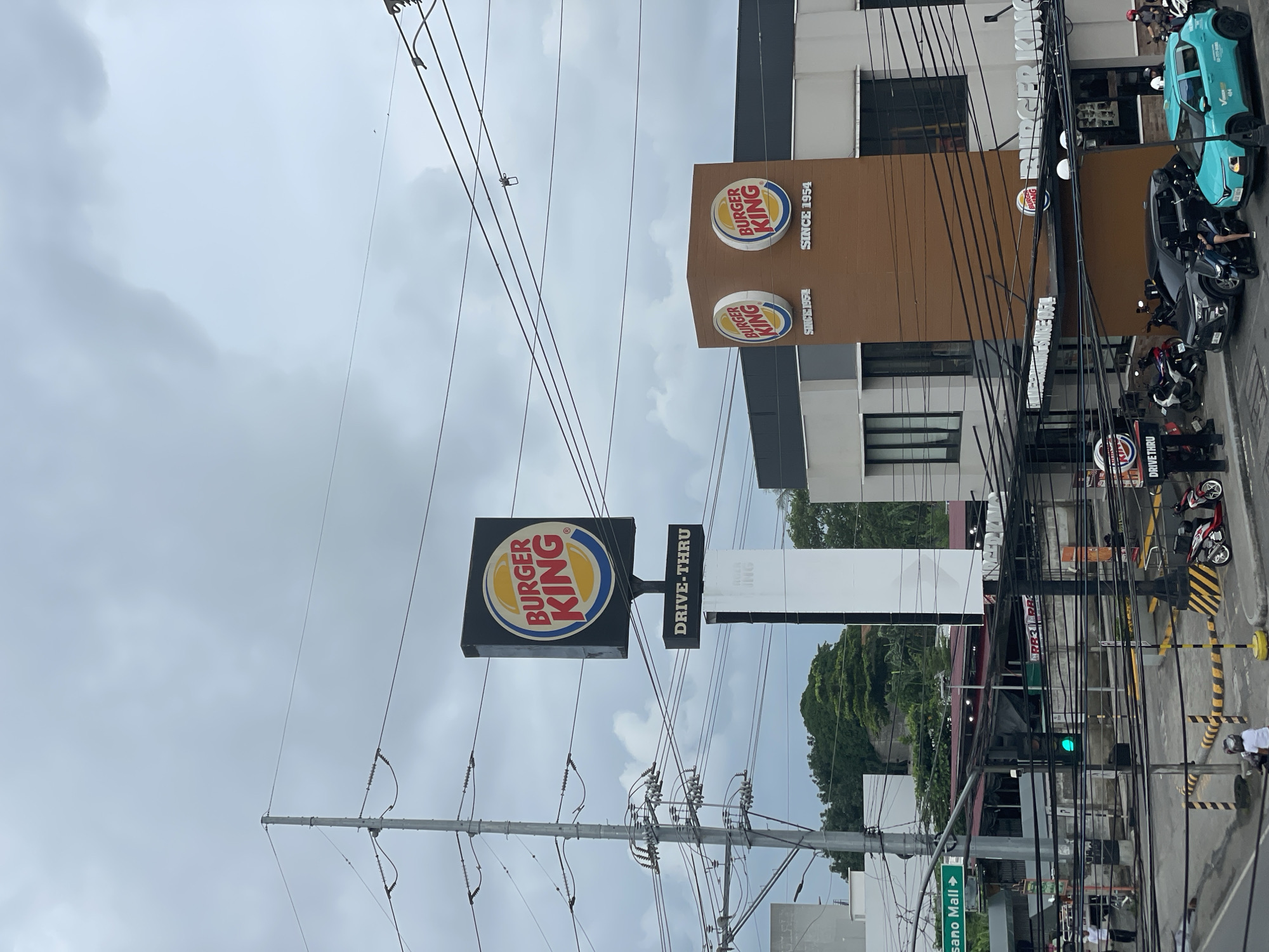 The image captures a Burger King restaurant in Davao City, Philippines, under an overcast sky. A large Burger King sign with the iconic logo is prominently displayed on a tall pole, accompanied by a "DRIVE-THRU" sign below it. A modern Burger King building with a brown facade and multiple Burger King logos is visible to the right. The scene includes a street with several vehicles, including cars, motorcycles, and a teal-colored car, parked or driving. Traffic lights are visible, with one showing a green light. Power lines crisscross the sky, characteristic of urban environments. Trees and other buildings can be seen in the background, suggesting a commercial or mixed-use area. The presence of the "DAVAO CITY, PHILIPPINES" text on a green directional sign confirms the location. The image appears to have been taken from a slightly elevated vantage point.