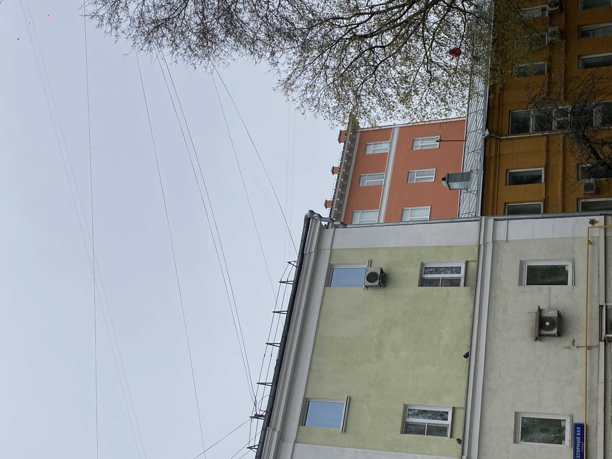 The image presents a tilted, upward-looking view of multi-story buildings and an overcast sky, with bare tree branches and utility wires crisscrossing the frame. The scene is set in an urban environment, specifically Moscow, Russia, as indicated by a visible street sign.
Three distinct buildings are prominent. On the left, a light green or pale yellow building with white window frames and multiple air conditioning units on its facade. To its right, a taller, beige or light grey building with similar windows and air conditioners. Further in the background, a vibrant orange-colored building with numerous windows is visible, partially obscured by other structures and tree branches. Another yellow-colored building is partially seen on the far right. The buildings feature various architectural details such as ledges, utility pipes, and railings.
Overhead, a pale, cloudy sky dominates the upper-left portion of the image. Numerous thin utility wires stretch across the sky from left to right. Tree branches, some with nascent green leaves, frame the top and right side of the image. A single red balloon is caught in the branches of a tree on the right side. In the far upper left, several small, indistinct pink or purple dots, possibly other balloons, are visible against the sky.
The time of day appears to be daytime, under an overcast sky, suggesting soft, diffuse light. There are no people visible, and no specific activities are taking place, presenting a quiet urban landscape. A blue street sign is visible on the lower right building, bearing the Cyrillic text "ЭГОРНЫЙ ВАЛ" (Egorny Val), with what appears to be "УЛ." (Ulitsa, meaning street) below it, confirming the Moscow location.