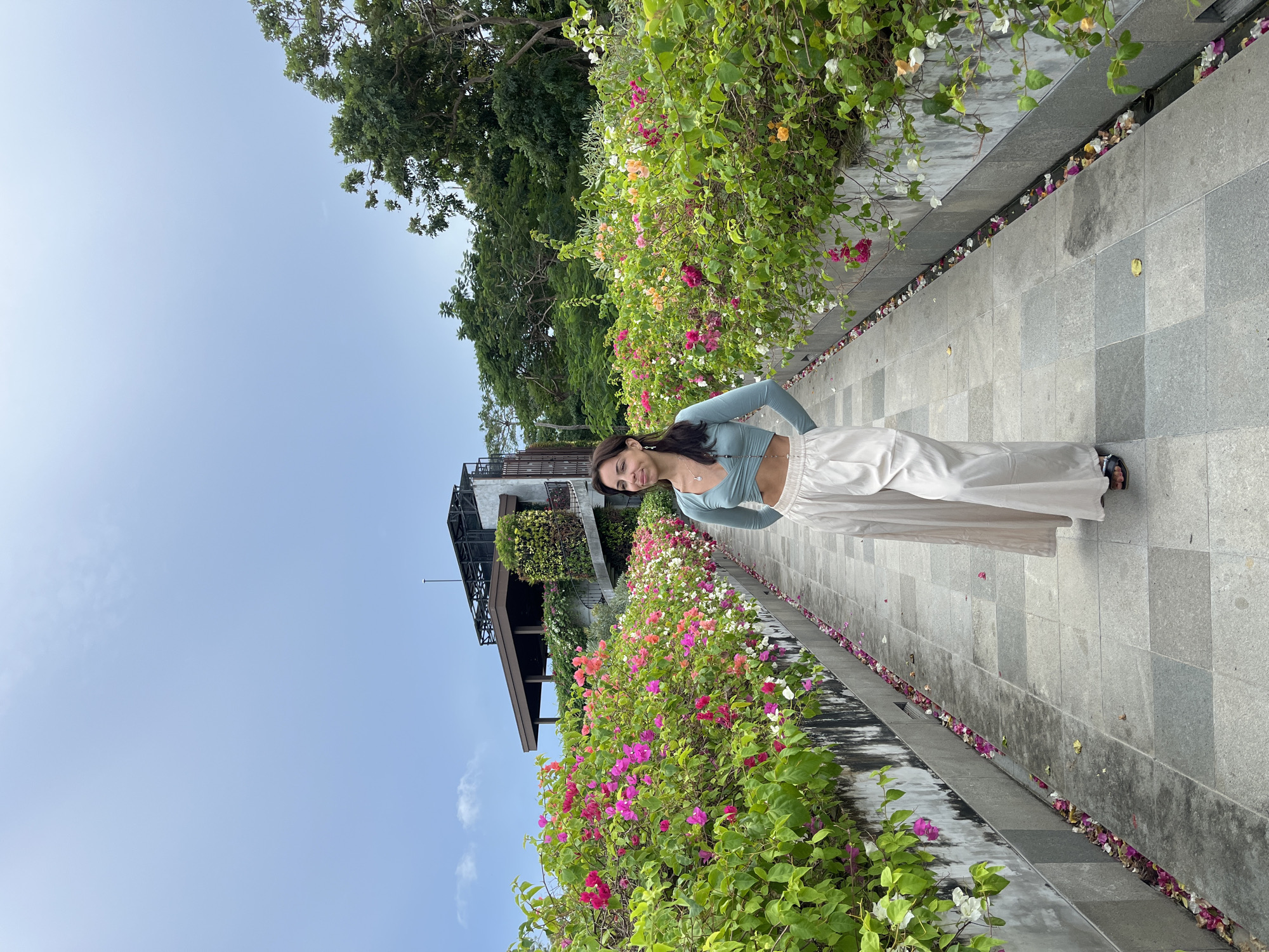 A woman is standing on a stone-tiled path in Kuta Selatan, Indonesia, posing for a photo. She is wearing a light blue long-sleeved crop top and loose, light-colored pants. Her hands are on her hips. Beside the path is a raised garden bed filled with colorful bougainvillea flowers in pink, white, and orange. Beyond the garden is a building with a dark roof and some greenery growing on its walls. The sky is a light blue. Petals of the flowers are scattered on the path next to the garden bed. The orientation of the photo is vertical.