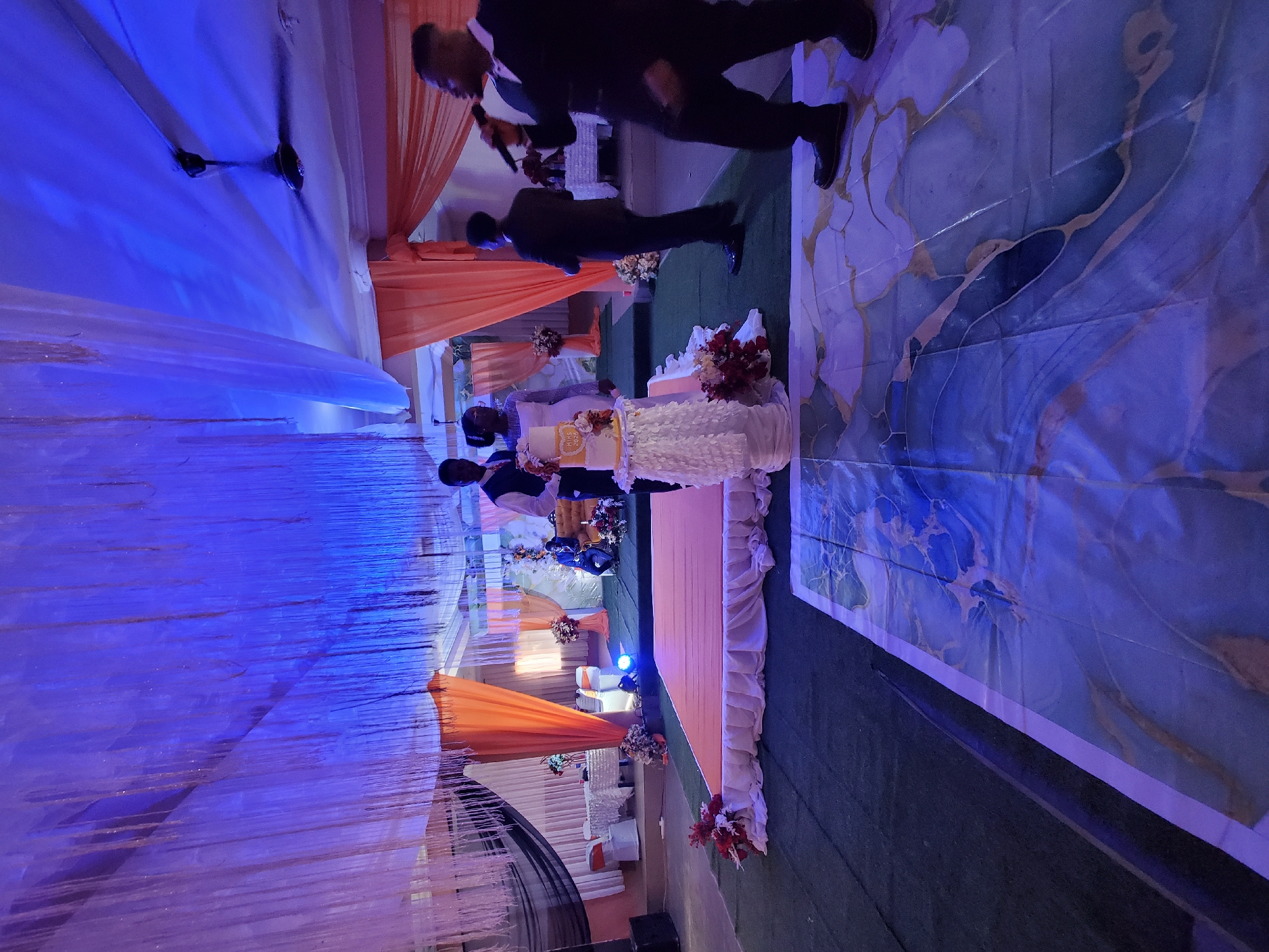 A wedding cake cutting is underway in a room in Onitsha, Nigeria. The scene, oriented vertically, is dominated by the layered white wedding cake adorned with flowers and gold accents on one of its layers. Two men stand behind the cake, presumably the bride and groom. Two men are standing on the far side of the image near the top, one holding a microphone. The room is decorated with white and orange drapes, and flower arrangements can be seen throughout. The floor is carpeted in green, and a large decorative rug, featuring blue and gold patterns, lies nearby. The lighting in the room casts a blue hue over the scene. Other tables, chairs, and decorations are visible in the background, completing the wedding reception atmosphere.