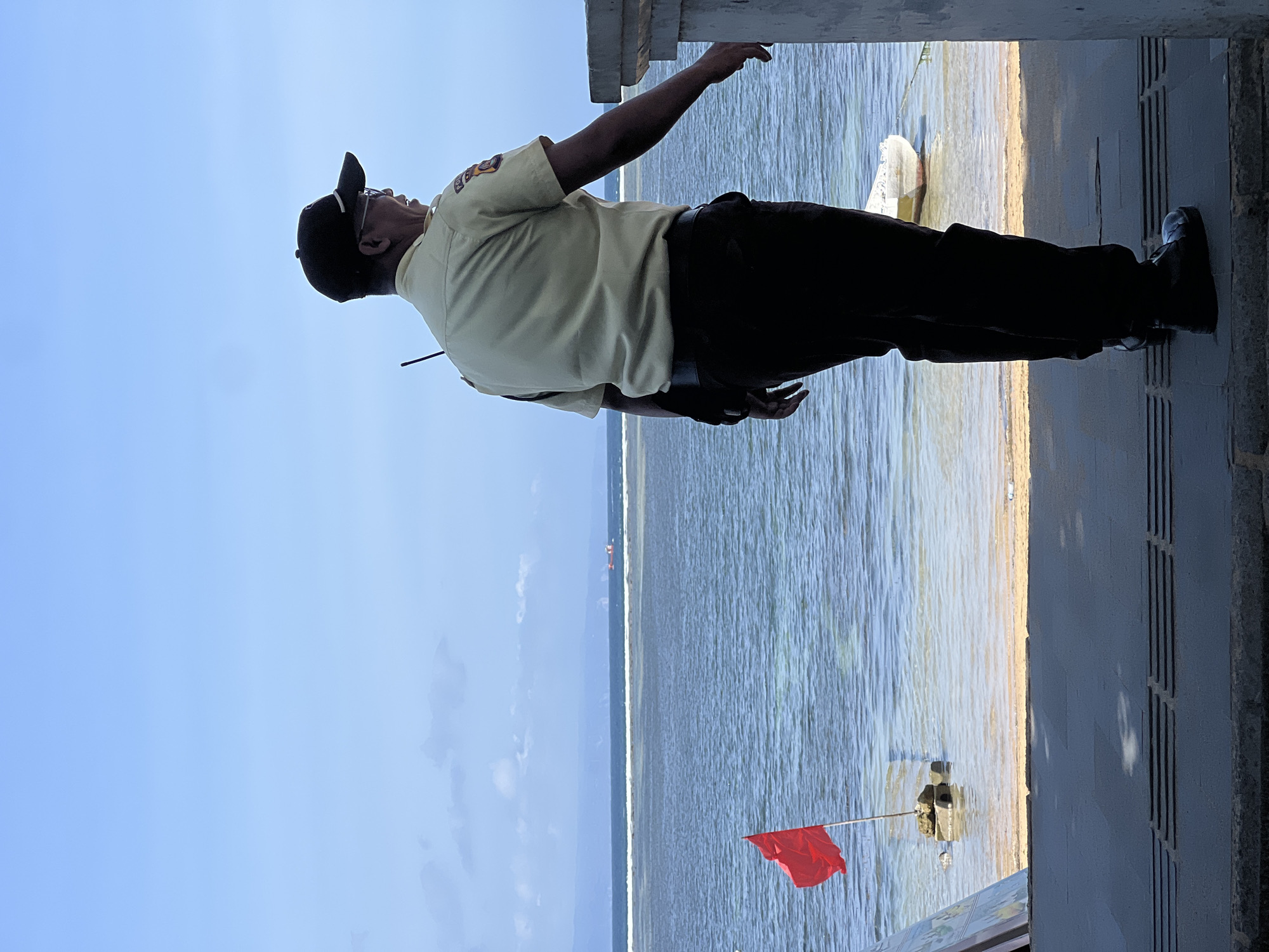 A man in a light-colored uniform and dark pants, wearing a baseball cap and glasses, stands facing away from the camera and looking out at the ocean. He has a radio clipped to his shirt. The scene appears to be a beach or coastal area. In the background, the ocean stretches to the horizon, with a few ships visible. A red flag is planted on a rock in the shallow water near the shore. The foreground is a dark platform with a textured surface. A white boat is moored in the water to the right of the frame. The sky is a pale blue with some scattered clouds. This scene is likely located on the coast of Denpasar, Indonesia, known for its beaches and maritime activity.