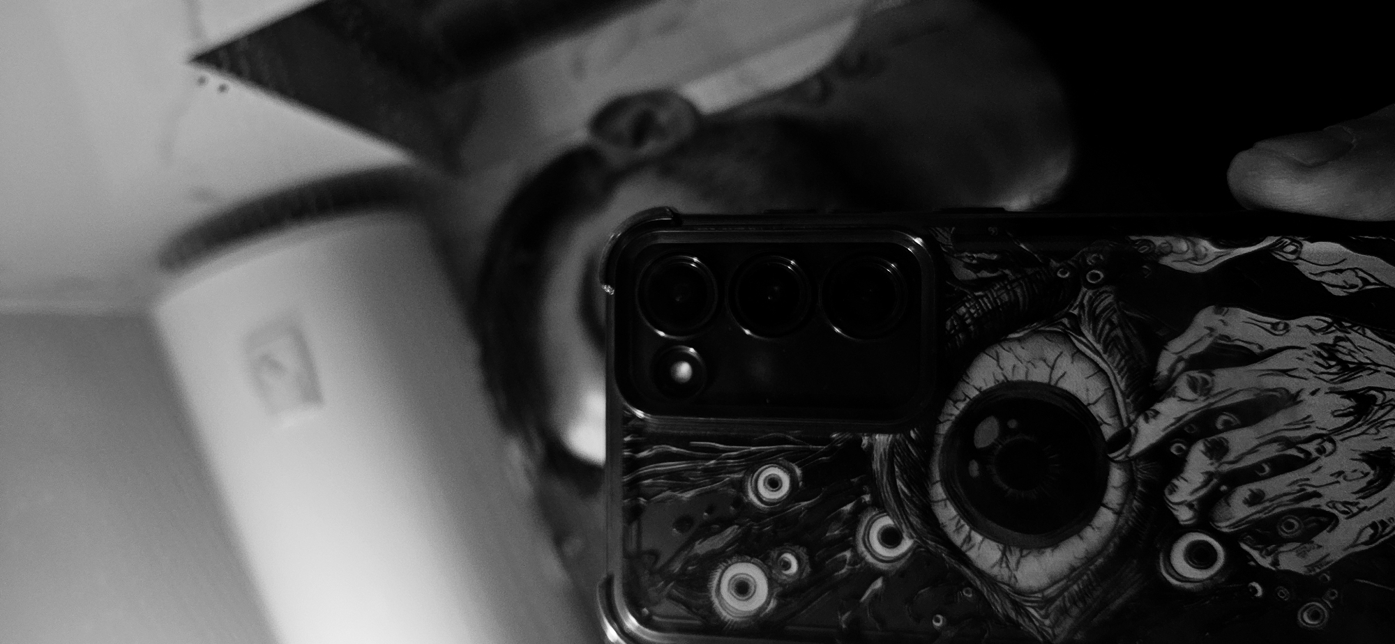 The image is a black and white, close-up photograph centered on the back of a smartphone. The phone is encased in a distinct dark-colored cover featuring intricate, macabre artwork. This design prominently displays a large, veiny eyeball on the right side, surrounded by multiple smaller eyes and several skeletal or claw-like hands reaching across. The phone's camera module, visible on the upper left of its back, shows three main lenses and a smaller sensor/flash.
The background is a blurred reflection on what appears to be a mirror or glossy surface. This reflection reveals the upper portion of a person's head, including an ear, and part of a shoulder. To the left of the reflected person, a blurry, light-colored object, possibly cylindrical or a section of a wall, with a small dark square feature, is discernible. Another dark, angular shape is reflected in the upper left corner of the background. The scene is dimly lit, contributing to the high contrast and atmospheric depth of the grayscale image. The primary activity depicted is the interaction with a smartphone, either being held or positioned for a reflective shot. There is no visible text within the image.