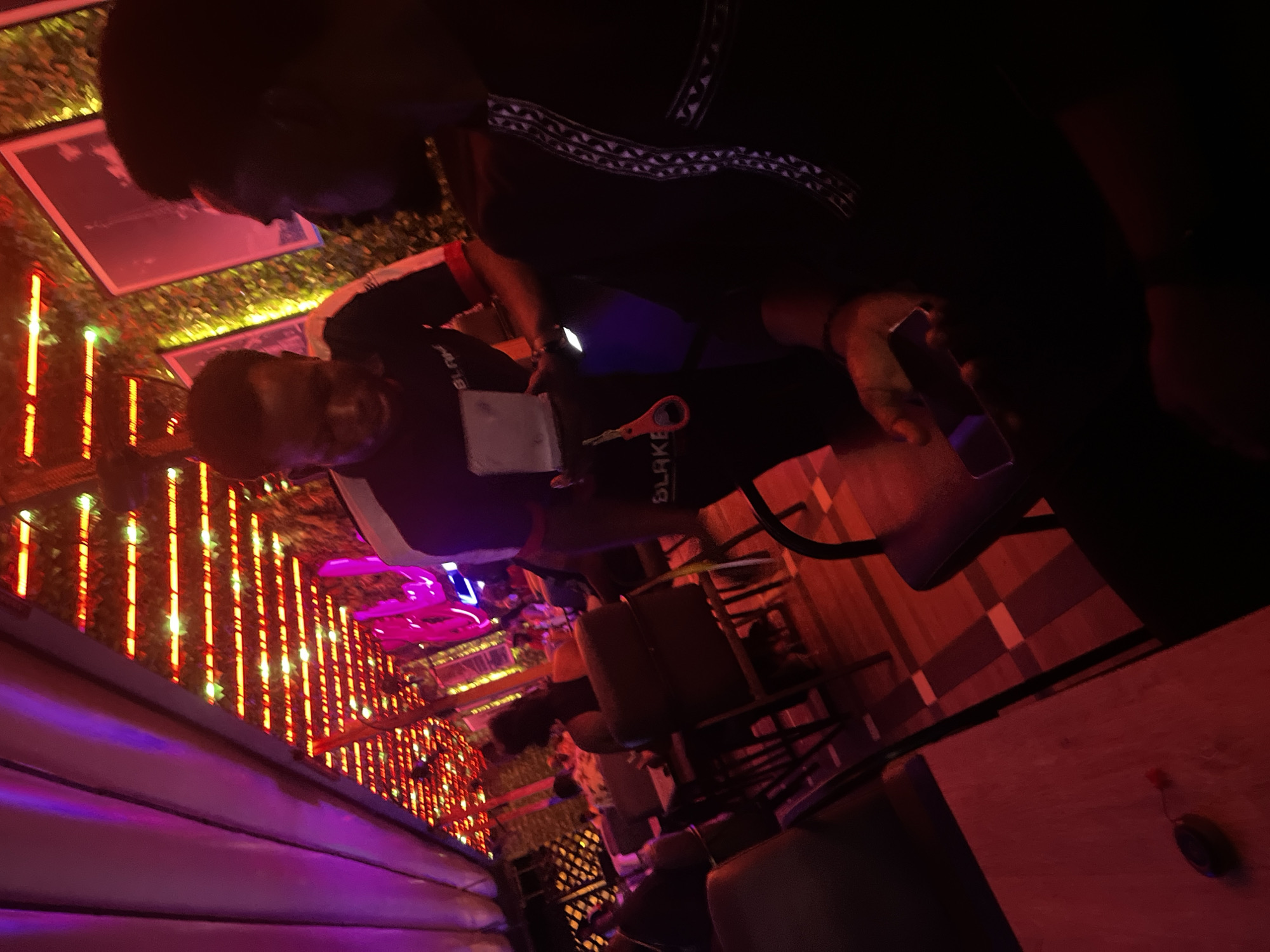 The image depicts an indoor establishment, identified as Blake Lounge & Cafe in Abuja, Nigeria, characterized by vibrant artificial lighting. The scene is illuminated predominantly by red-orange LED strip lights arranged in parallel rows across the ceiling and purple ambient lighting from various sources, including what appears to be decorative panels or neon signs.
In the mid-ground left, a male staff member is standing, facing towards the right. He is wearing a dark short-sleeved shirt with the word "BLAKE" visible on the chest, along with red and white trim on the sleeves. He holds a small, white rectangular object, possibly a menu or tablet, in his left hand, and a red, key-like object is attached to his left hip area.
To the right, in the foreground, a male patron is seated, seen from his right side. He is wearing dark clothing with a distinct white and black geometric pattern running vertically down the side of his garment. He holds a smartphone horizontally in his hands; its screen emits a faint purple glow.
Further in the background, several other patrons are seated at tables, their forms largely indistinct due to low light and focus. The establishment features dark upholstered seating and wooden tables. The flooring is a light-colored wood or laminate with darker rectangular sections creating a patterned effect. Walls are adorned with artificial green foliage and framed pictures or artwork. A small, dark, ovular object with a red attachment, possibly a key fob, rests on a table in the bottom-left foreground.