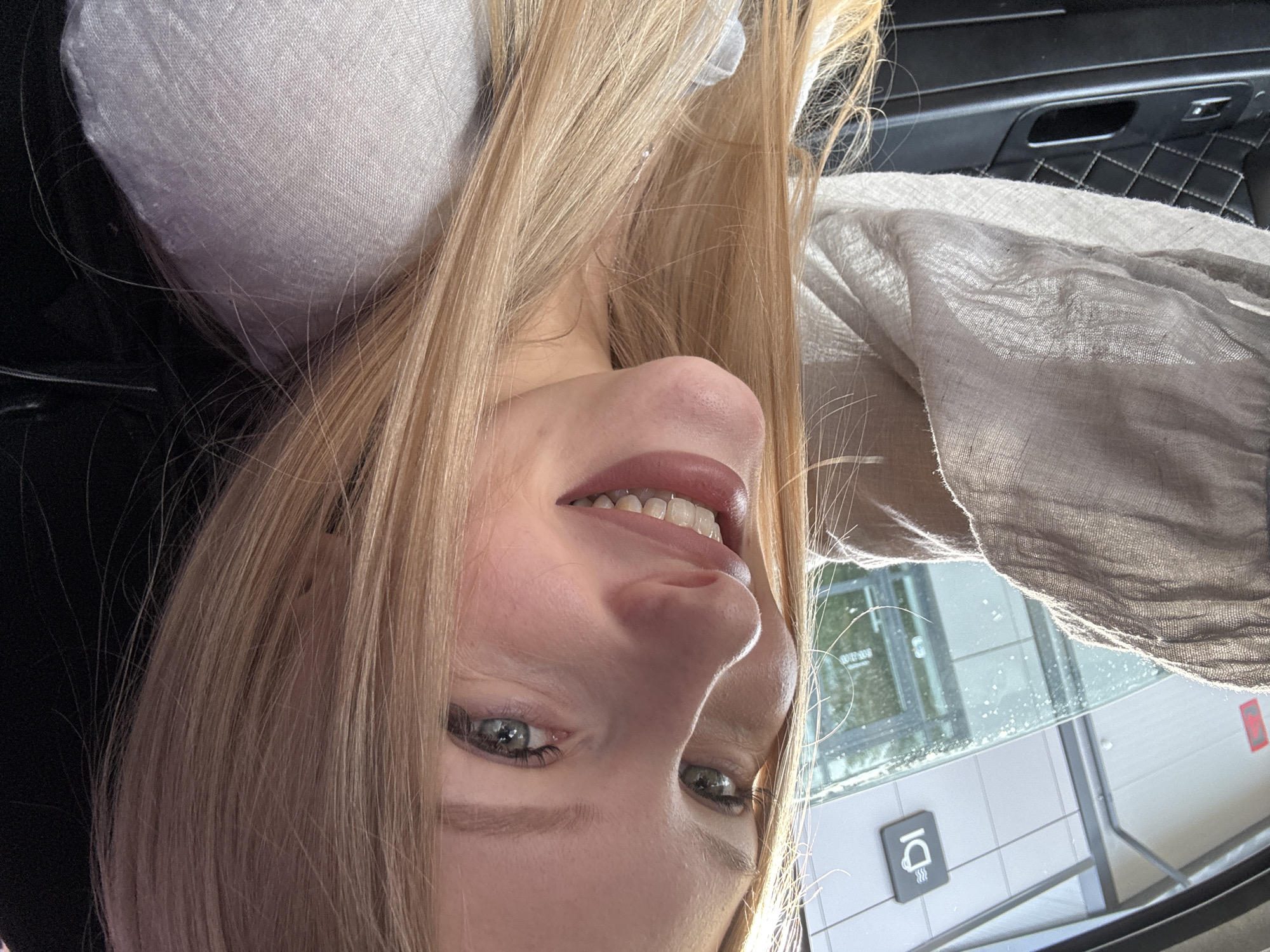 A woman with blonde hair and blue-green eyes is taking a selfie in a car. She is wearing a light-colored shirt. Through the car window, a building with a coffee cup logo is visible, suggesting a cafe. The scene appears to be in the city of Perm, Russia, indicated by the context of the request. The woman is smiling at the camera. The car's interior is partially visible, showing a black upholstered door panel with diamond stitching and a window control. The lighting suggests it is daytime, possibly with sunlight filtering through the window.