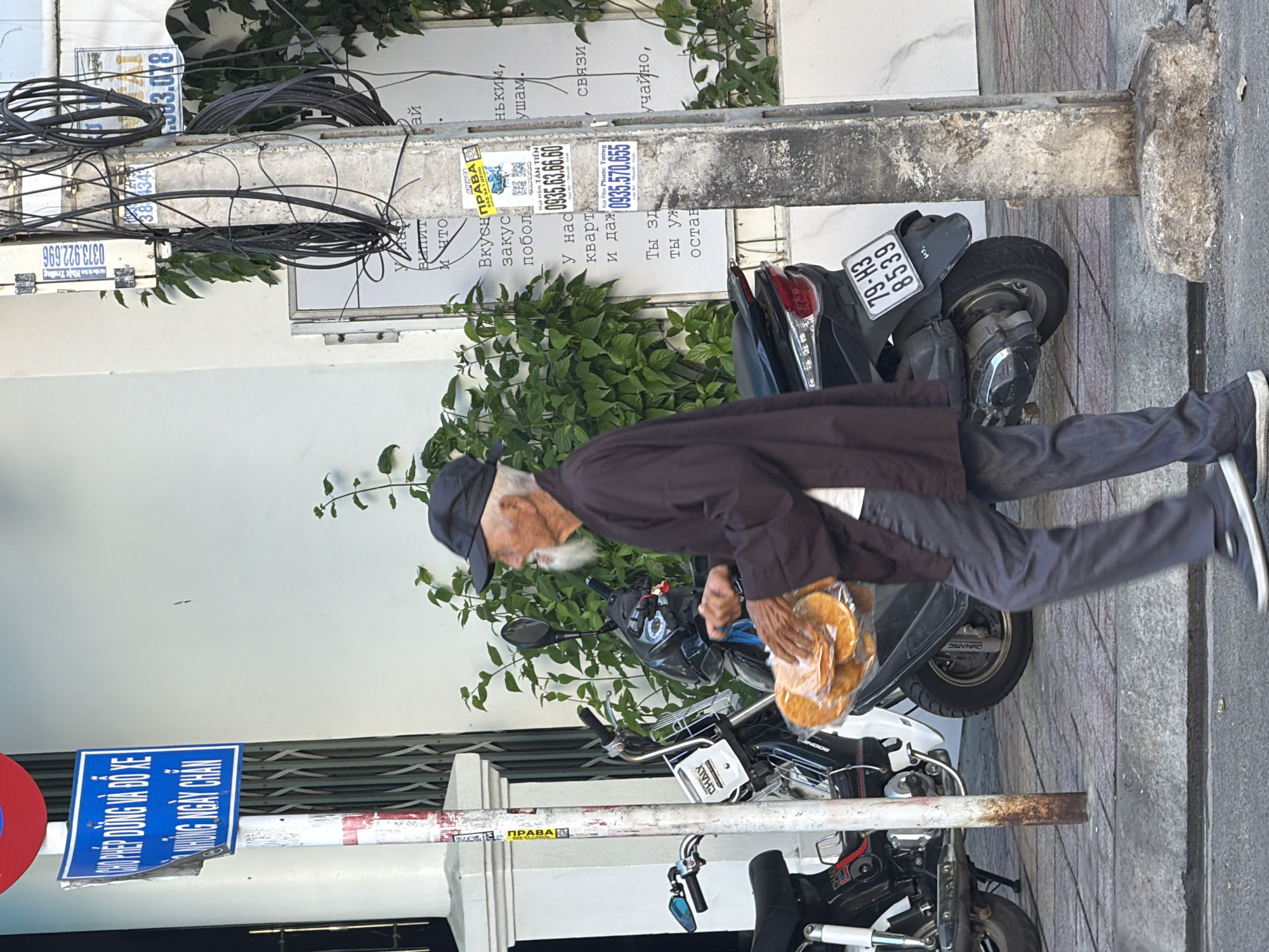 An elderly Vietnamese man with a white beard, wearing a black baseball cap and a dark brown tunic over gray pants, walks from left to right across a sidewalk in Bac Nha Trang Ward, Vietnam. He carries a clear plastic bag filled with round, flat food items, possibly bread or crackers, in his right hand. He is walking past two parked motorcycles, a white one on the left and a black one on the right. A utility pole with numerous black wires and several stickers is located to the right of the man. A blue and white street sign is visible on a pole behind the man to the left, with Vietnamese text. The ground is paved with cobblestones, and a curb separates the sidewalk from the street in the foreground. The background features a white wall and green foliage.