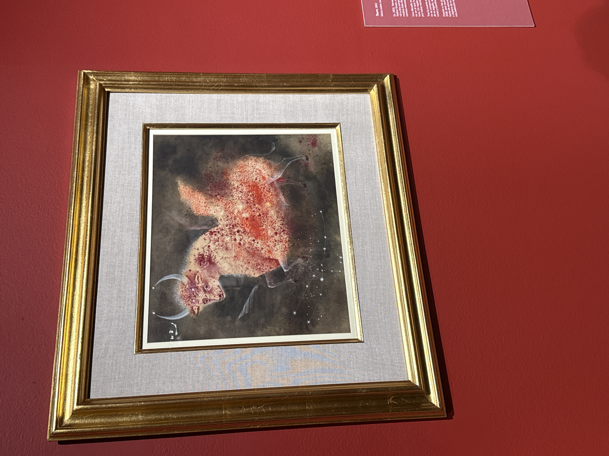 The image shows a framed painting of a bull on a red wall. The painting, matted in gray fabric, is encased in a gold-colored frame. The bull is painted in shades of red and white, and it has a constellation of stars above it. The painting's title is "Tauro, 1962," and it is described as "Mixta sobre cam." The wall is red. Based on the included OCR, the painting is located in Mexico City, Mexico.