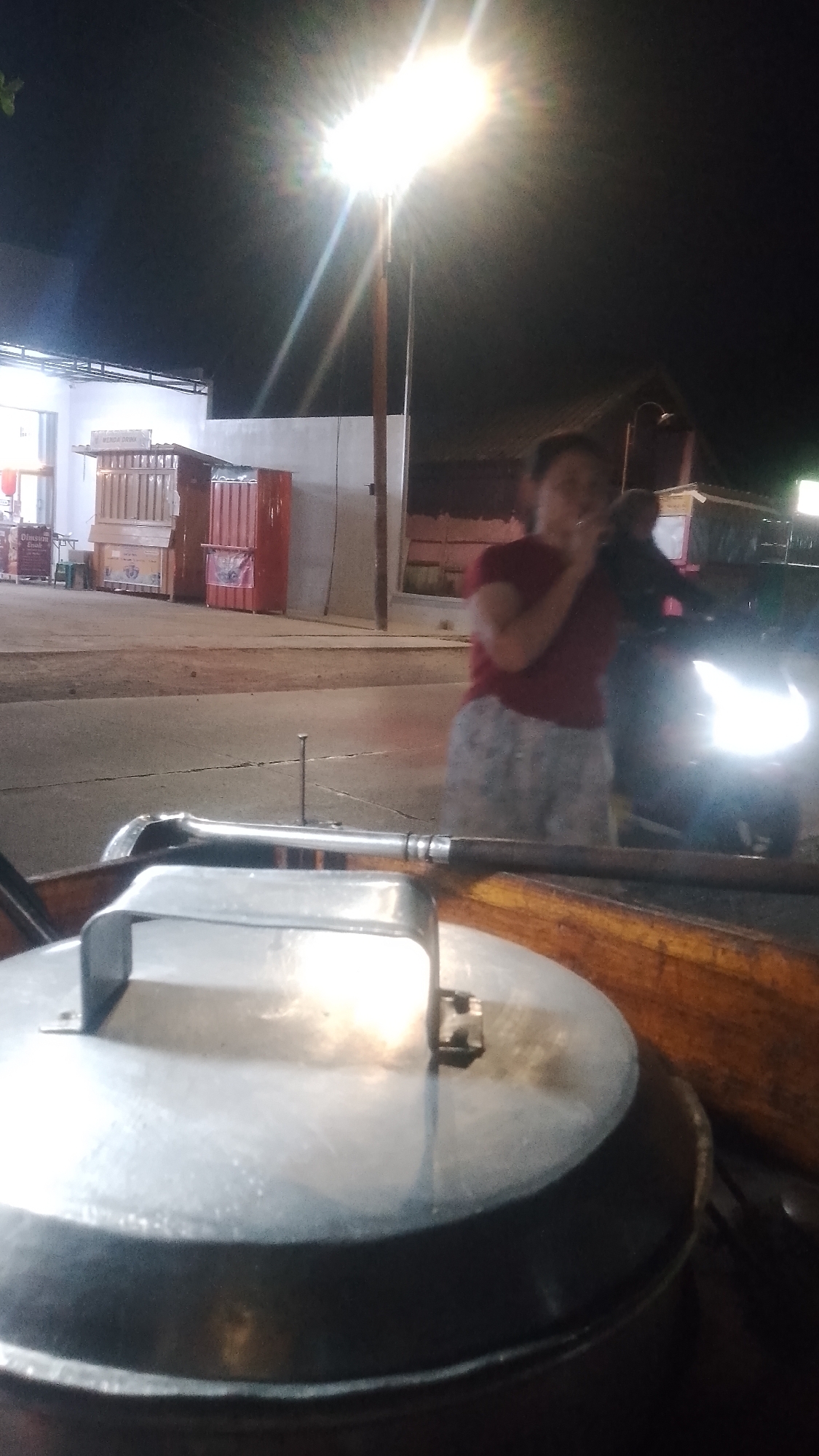 At night in the city of Karanglangu, Indonesia, an outdoor scene unfolds. A bright streetlamp illuminates the area, casting stark shadows. In the foreground, the lid of a large metal pot is visible, its handle indicating it's part of a food stall or cart. In the background, a woman wearing a red shirt and patterned shorts stands near a motorcycle, seemingly smoking. The woman is the primary human subject. Other elements in the environment include various food stalls or small shops with colorful structures, and a paved road. The overall atmosphere is that of a typical Indonesian night market or roadside food vendors.
