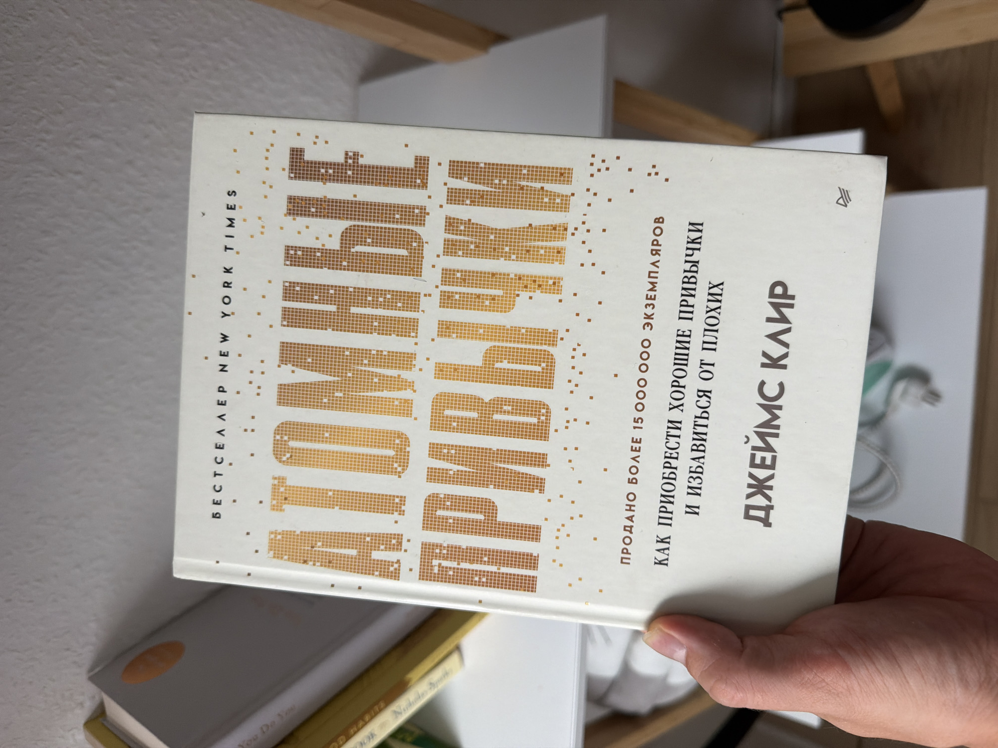 The image shows a hand holding a book with the title "Atomic Habits" in Russian. The book is white with gold lettering. The title is in large, bold letters, with smaller text below. The background is blurred, but appears to be a shelf with other books and a light-colored wall. There is no indication of the city of Herne, Germany in the image.