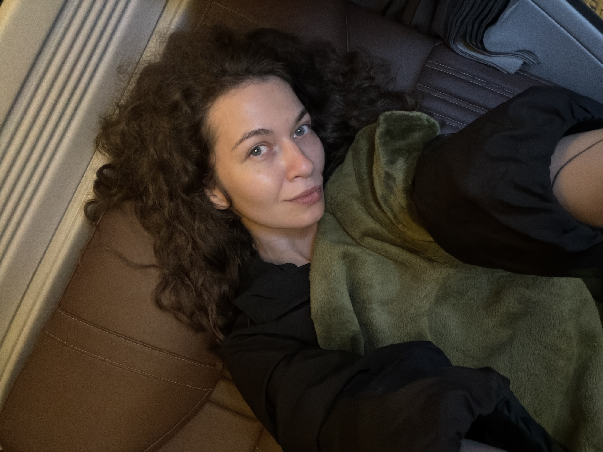 A medium-close-up shot shows a woman with curly dark brown hair and light eyes reclining in what appears to be the backseat of a vehicle or on a sofa. She gazes directly at the camera with a calm expression. Her hair is spread out around her head, resting on brown, possibly leather, upholstery with distinctive white stitching. She is wearing a black long-sleeved top and is partially covered by a soft, olive-green blanket or throw.
In the upper left background, vertical parallel lines are visible, suggesting blinds or a textured panel. On the right side, a grey folded fabric item rests on the upholstery. The overall lighting is soft, creating a relaxed and intimate atmosphere. The setting suggests a moment of rest or travel, possibly in a car in Đà Nẵng, Vietnam. No text is visible in the image.