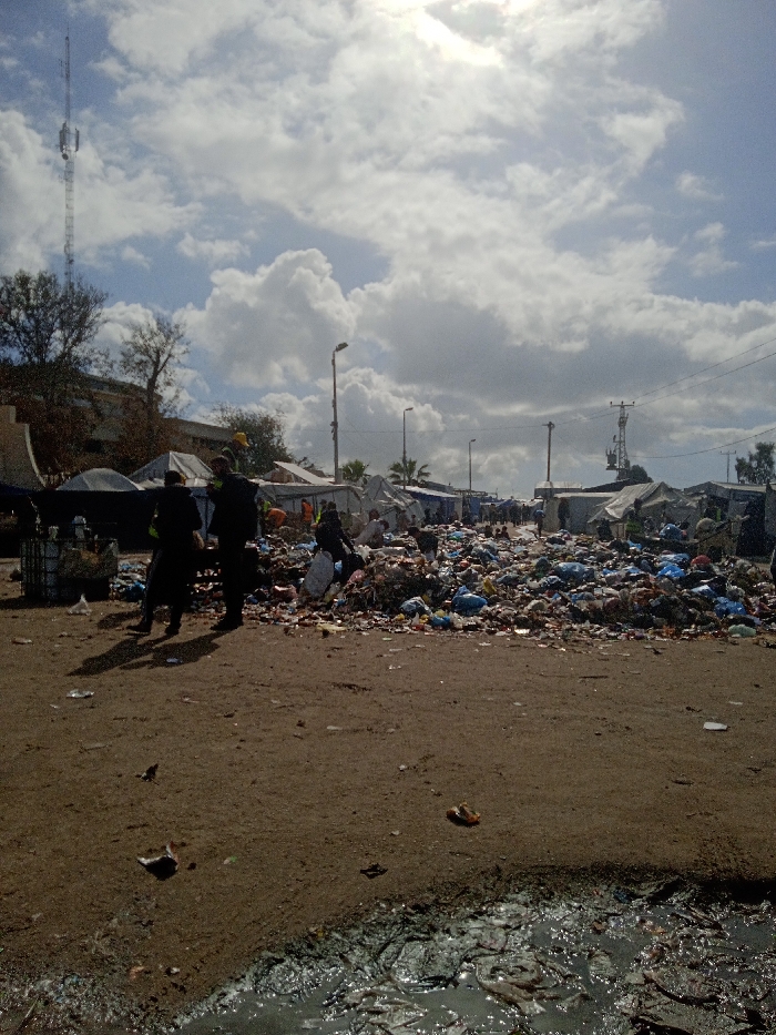 The image depicts an outdoor scene in Khan Yunis, Palestinian Territory, characterized by a makeshift settlement and pervasive refuse under a partly cloudy sky with bright sunlight. In the immediate foreground, a dark, murky puddle of liquid mixed with debris, likely wastewater, is visible on a light brown dirt surface scattered with litter.
Dominating the midground is a large, elongated mound of accumulated garbage, consisting of numerous plastic bags, discarded packaging, and various waste items, extending horizontally across the frame. Two individuals in dark clothing are seen walking from left to right near the left edge of this refuse pile. Another figure is observed bent over within the garbage further to the right. Additional indistinct individuals are present deeper within the scene, near the tents. To the left of the garbage, a wire mesh container or bin is partially visible, also surrounded by refuse. A line of utility poles with overhead power lines stretches across the midground.
The background features a dense encampment of numerous makeshift tents and shelters, constructed from light-colored tarpaulins, extending into the distance on both sides of a central pathway. On the far left, a multi-story building is partially visible behind trees and tents, next to a tall, lattice-structured communications tower. Another smaller tower is present on the right side of the background. Scattered trees are visible throughout the background. The sky is mostly light blue with large, white cumulus clouds, and a bright glare from the sun is present in the upper center.