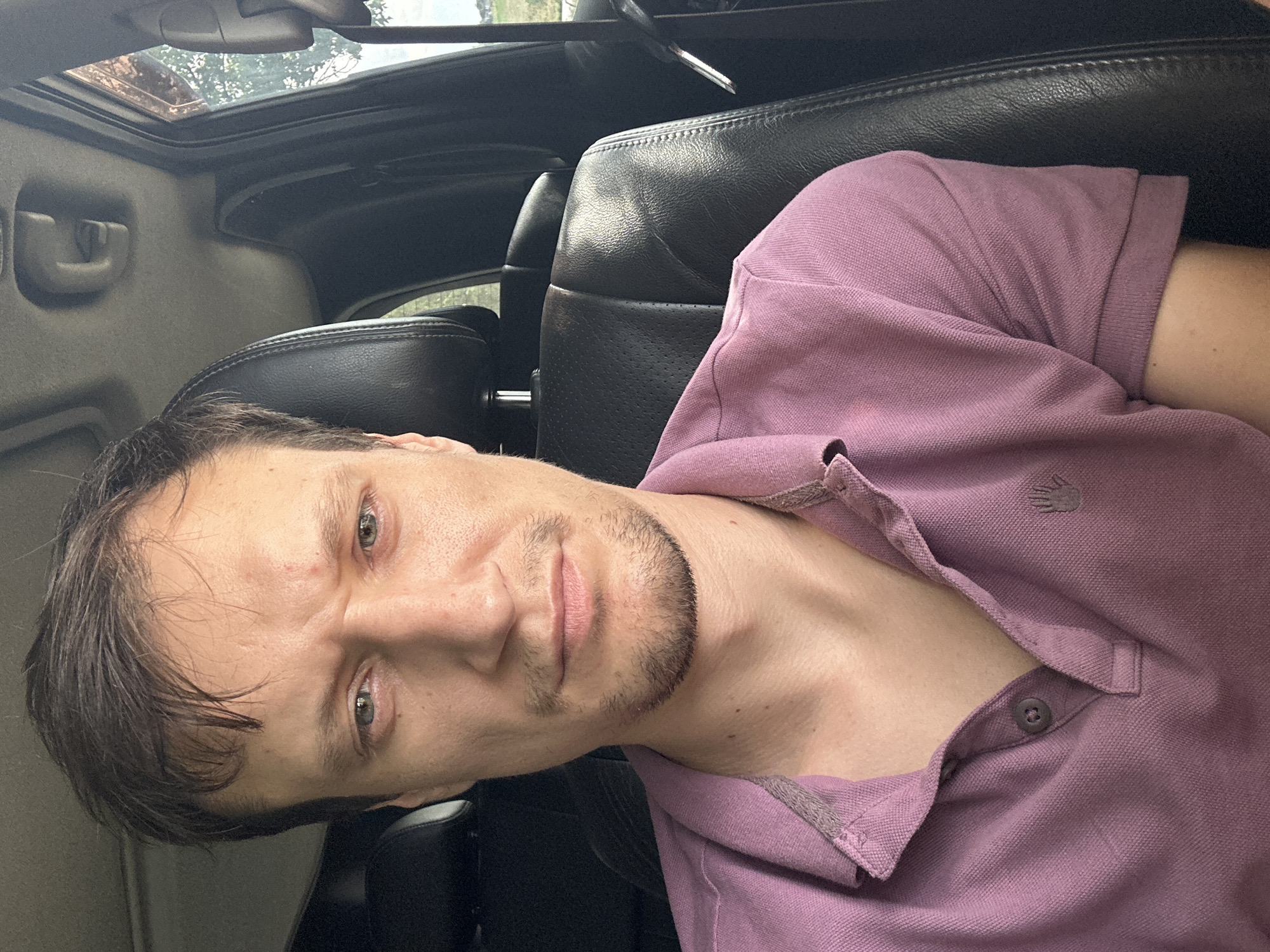 A close-up, selfie-style shot shows a fair-skinned man with short, dark brown hair and a light beard. He is wearing a pink polo shirt with a collar and two buttons. He is seated in the driver's seat of a vehicle, as indicated by the car's interior visible in the background. The black leather headrest and seat are prominent. The man's facial expression appears serious or concerned. The lighting suggests it is daytime, and the outdoor environment is visible through the car window on the right, with trees and a hint of sky. No specific landmarks of Makiv, Ukraine, are identifiable within the image.