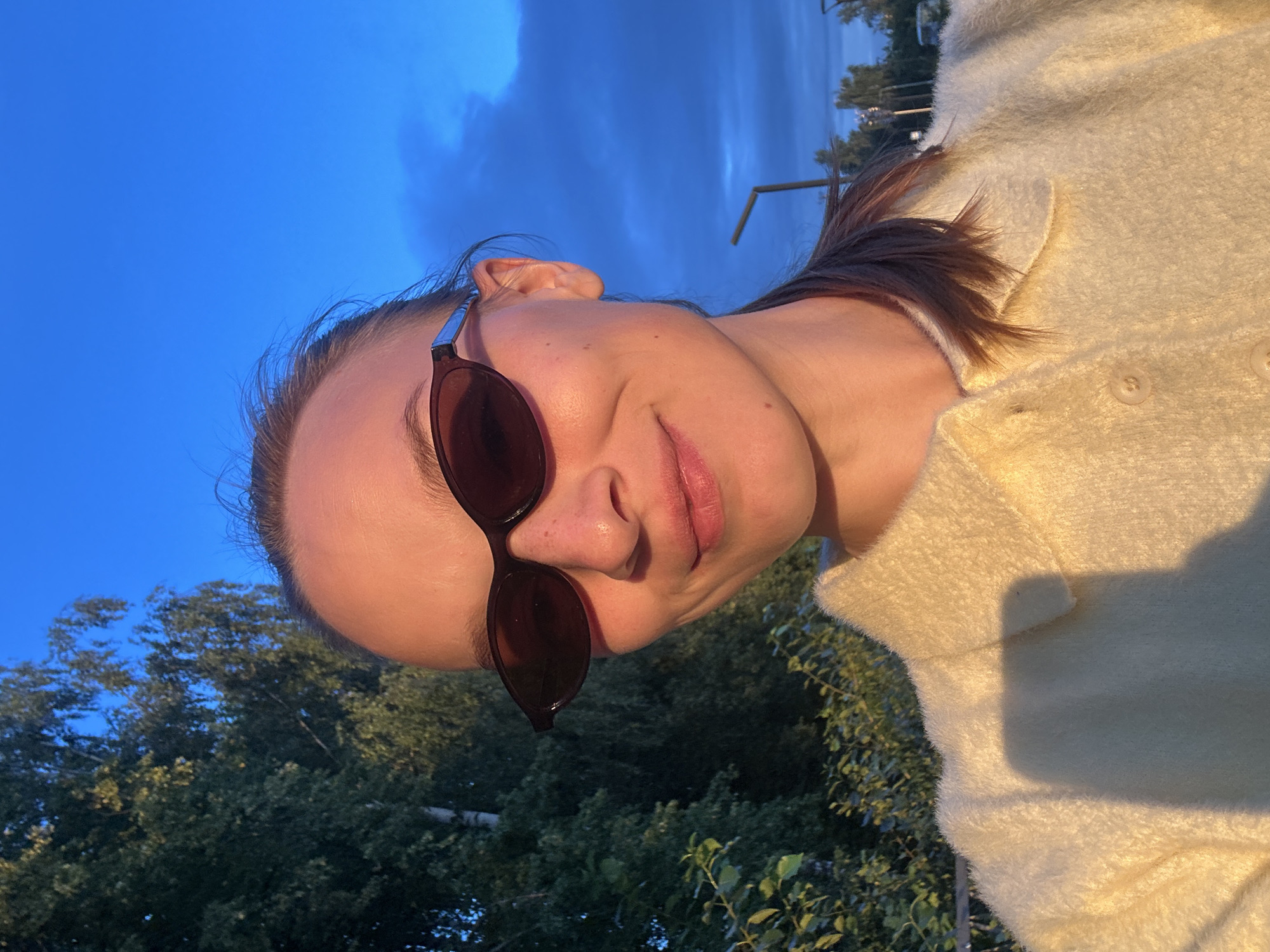 A young woman is taking a selfie outdoors. She has blonde hair pulled back in a ponytail, and she is wearing large, dark sunglasses and a light yellow fuzzy collared shirt. She is smiling at the camera. Behind her, there are green trees and a bright blue sky with some clouds. In the background, there are also some streetlights and more trees. The lighting suggests it is either early morning or late afternoon. The location is in the city of Chaikovsky, Russia.