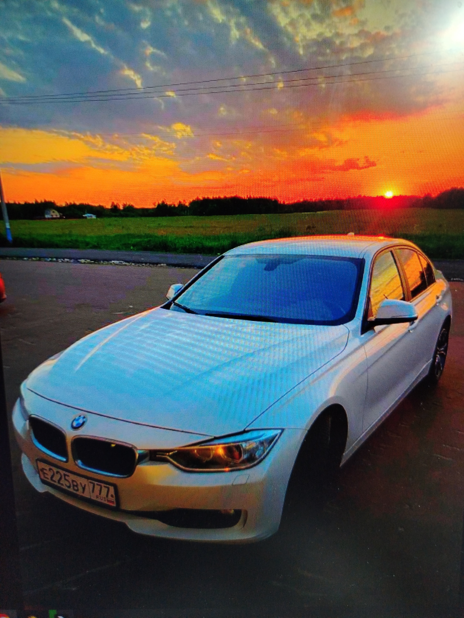 The image features a white BMW sedan parked on a paved surface with a sunset in the background. The car has a Russian license plate that reads "E225BY 777". The background shows a field and some trees under a vibrant orange and yellow sunset. There are power lines visible in the upper part of the frame. The location appears to be outside a city, given the presence of fields and trees. The reflections on the car's surface show the colors of the sunset.