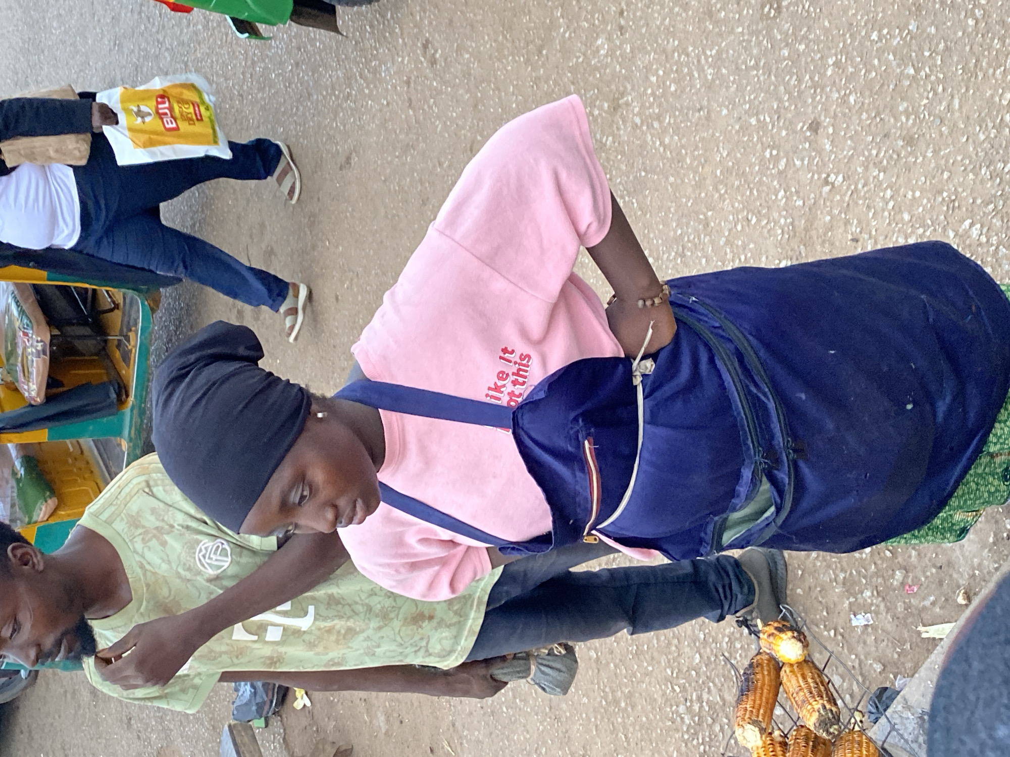 In Bukuru, Nigeria, a young woman wearing a black headscarf and a pink t-shirt with red lettering that says "like it ot this" is standing on a dirt road. She is also wearing a dark blue apron over a green patterned skirt. Her hands are on her hips. To her left, a man with a beard, wearing a light green t-shirt, is standing. He is holding a small, wrapped object in his left hand. In the background, a yellow and green motorized rickshaw is visible. Roasted corn on a grill is in the lower left of the frame. A person in the background is carrying a yellow "BULL" branded bag.