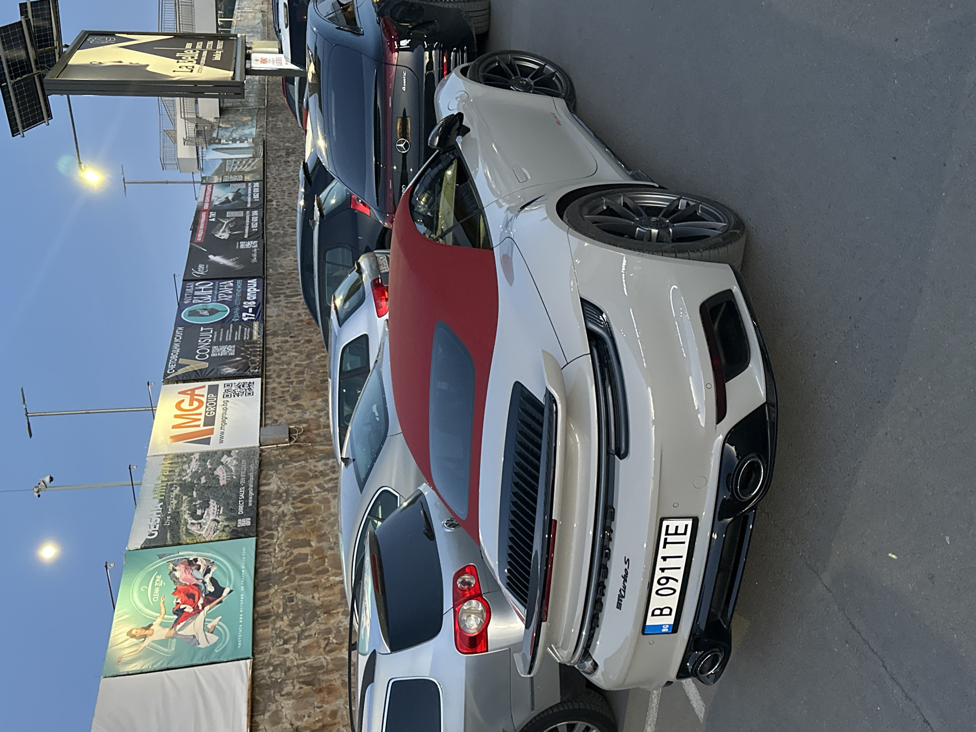 The image captures a parking area in Varna, Bulgaria, identifiable by the vehicle registration plate "B 0911 TE" indicating a Bulgarian registration and signs with Bulgarian text in the background. The focus is on a white Porsche 911 Turbo S with a red convertible top. It's parked alongside several other cars, including a black Mercedes-Benz 4MATIC. In the background, there is a wall with multiple advertisements, including one for "La Belle" and another for "CLEAN ZONE." One sign indicates "Gesha Hillpark" and another advertises financial services ("СЧЕТОВОДНИ УСЛУГИ"). A festival advertisement shows "ХРАНА" and the dates 17-18 април, implying a food festival in April. A sign for "MGA GROUP" is also visible. Street lights are present, indicating the time of day could be dusk or evening. There are no people visible in the image.