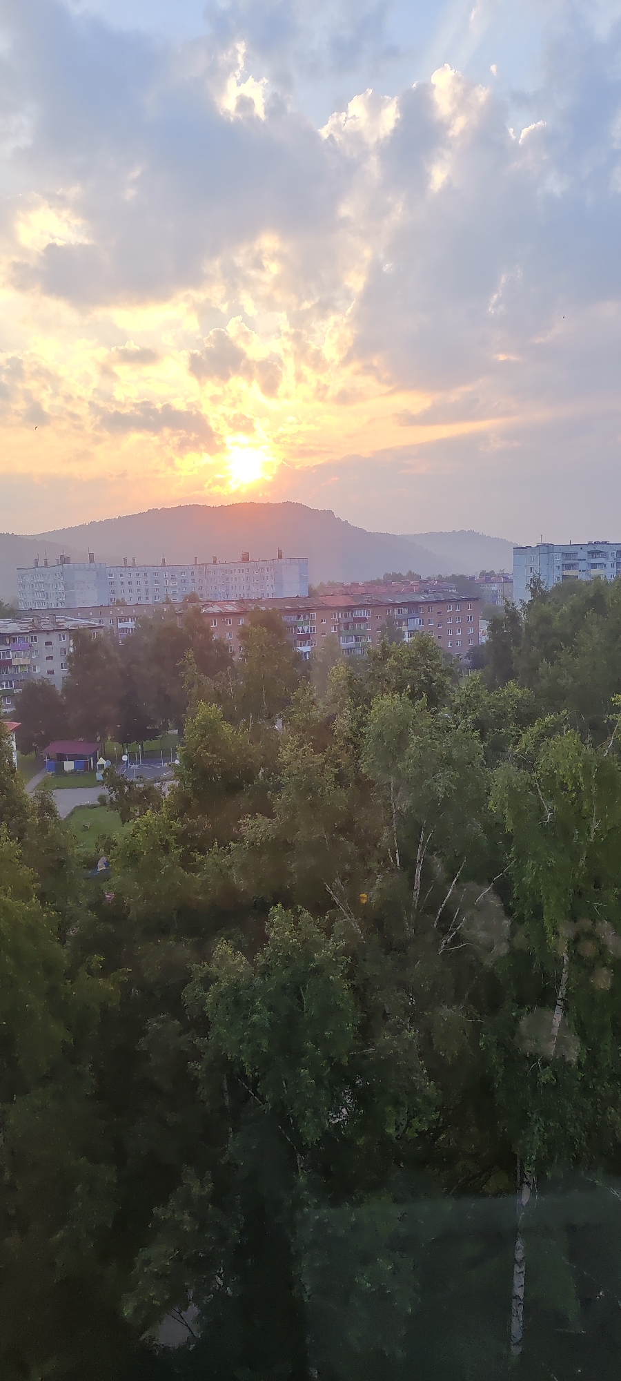 The image shows a sunset over a city. The sun is visible in the sky, casting a golden light. There are clouds in the sky, some of which are illuminated by the sun. In the background, there are hills. In the middle ground, there are buildings, some of which are tall apartment buildings. In the foreground, there are trees. The scene appears to be a residential area, with apartment buildings and trees visible. The city is Mezhdurechensk, Russia.