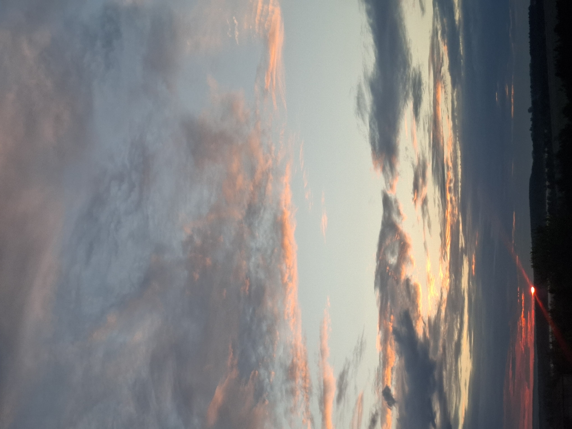 The image depicts a sunset over a body of water, likely a lake or river, possibly located near Khlevnoye, Russia. The sky is filled with clouds, some illuminated with orange and pink hues from the setting sun. The water reflects the colors of the sky. Silhouetted trees line the distant shore.