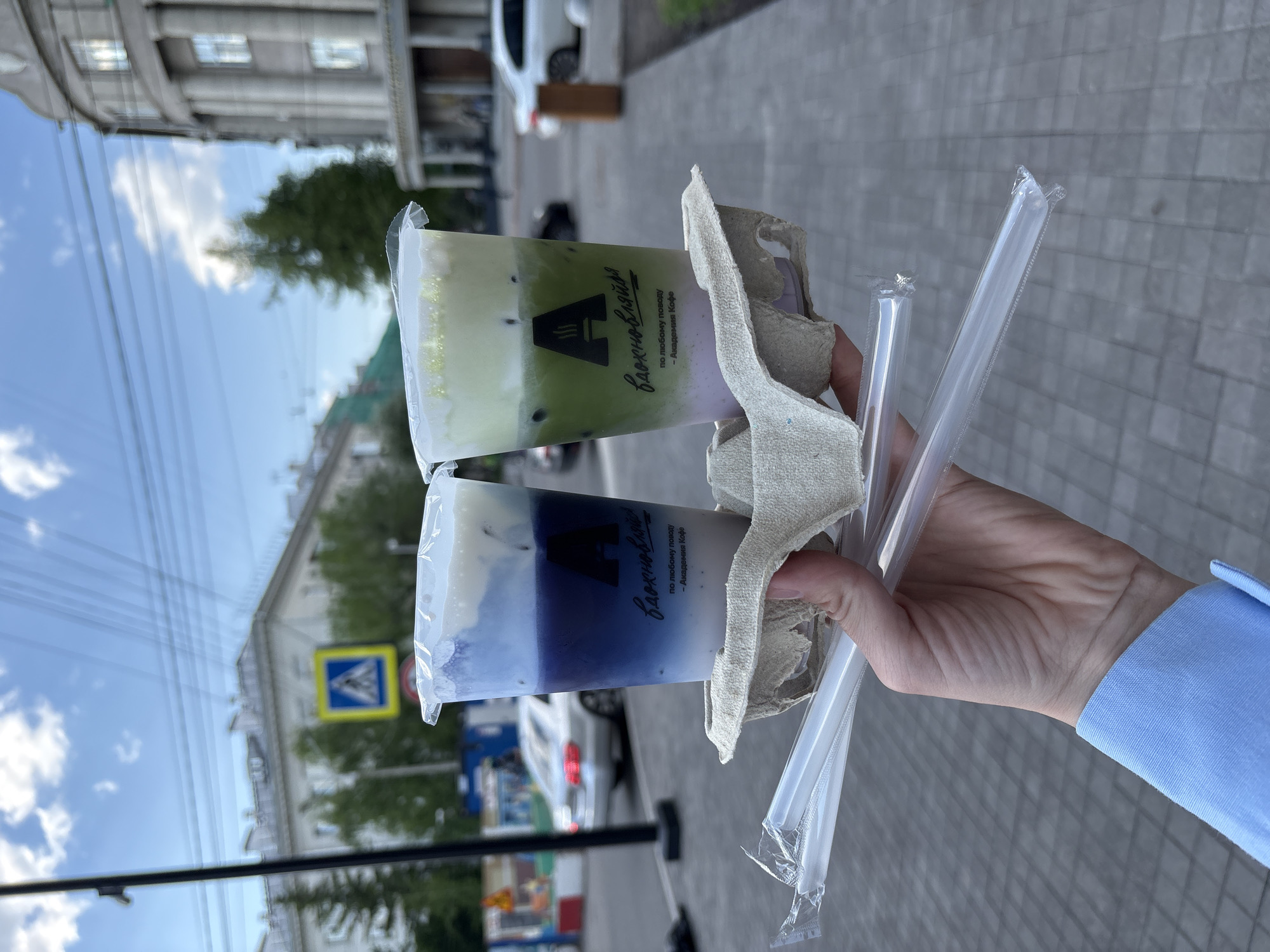 A hand holds a beverage carrier with two iced drinks in plastic cups, each sealed with plastic wrap and containing straws. One drink has a white top layer over a green layer with dark specks, and a purple-tinted bottom layer. The other drink has a white top layer over a blue-to-purple gradient layer. Both cups have a logo with a stylized letter "A" and the words "Вдохновляйся" and "по любому поводу - Академия Кофе" in Cyrillic script. The carrier is made of a light-colored, possibly recycled material. The hand is wearing a blue sleeve. The background shows a city street with buildings, trees, cars, and a crosswalk sign. The location appears to be Krasnoyarsk, Russia, based on the Cyrillic script on the cups.