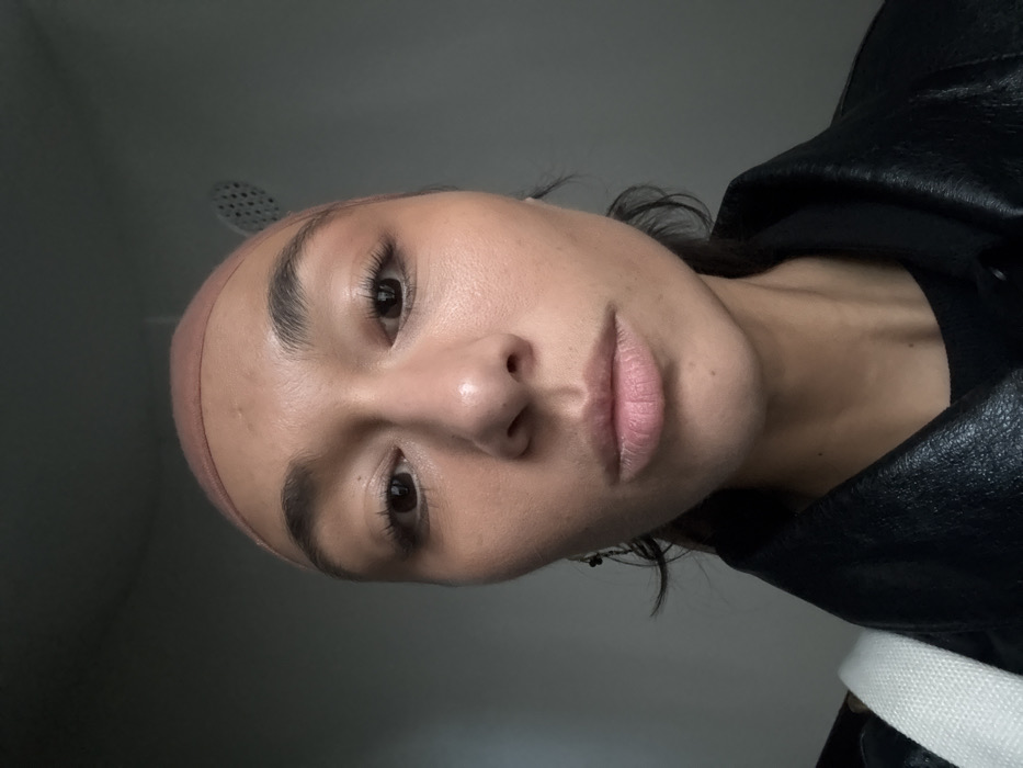 A close-up, front-facing photographic portrait depicts an adult female with a neutral expression. She has a medium skin tone, dark eyes, full eyebrows, and her lips are closed. Her head is covered by a light brown, form-fitting cap. She is wearing a black jacket, likely leather, over a dark undergarment. A dark earring is visible on her left ear, and a white, textured strap, possibly from a bag, is visible over her left shoulder. The background is a plain, light gray wall, with a partially visible circular, perforated object, consistent with a showerhead or ventilation grate, in the upper right. The scene is indoors, within what appears to be a bathroom or shower enclosure. The image provides no specific visual cues to identify a precise location within the city of Paris, France.
