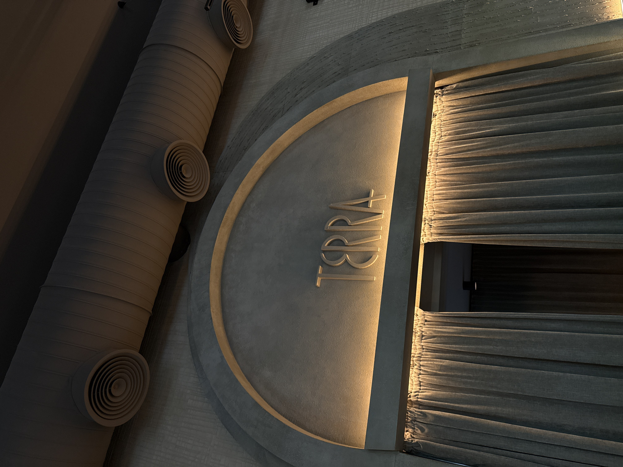 This image captures an interior architectural detail, likely within an upscale commercial establishment like a restaurant or lounge in Yerevan, Armenia. The main subject is a large, circular sign made of a light-colored, textured material, possibly concrete or stone. It features a prominent, warm golden back-lighting that illuminates its edges and the embossed, stylized text "TERRA" in all caps.
To the left of the sign, a large, dark grey or beige corrugated ventilation duct runs diagonally, fitted with at least three circular air vents. To the right, a dark grey, heavily pleated curtain hangs vertically, also illuminated by a warm light source, suggesting an entrance or window. The overall aesthetic is modern and sophisticated, characterized by muted grey tones and strategic artificial lighting. The perspective is an angled overhead shot, focusing on the interplay of these design elements. No people or specific activities are depicted, emphasizing the architectural details. The lighting suggests an indoor setting, likely in the evening or at night.
