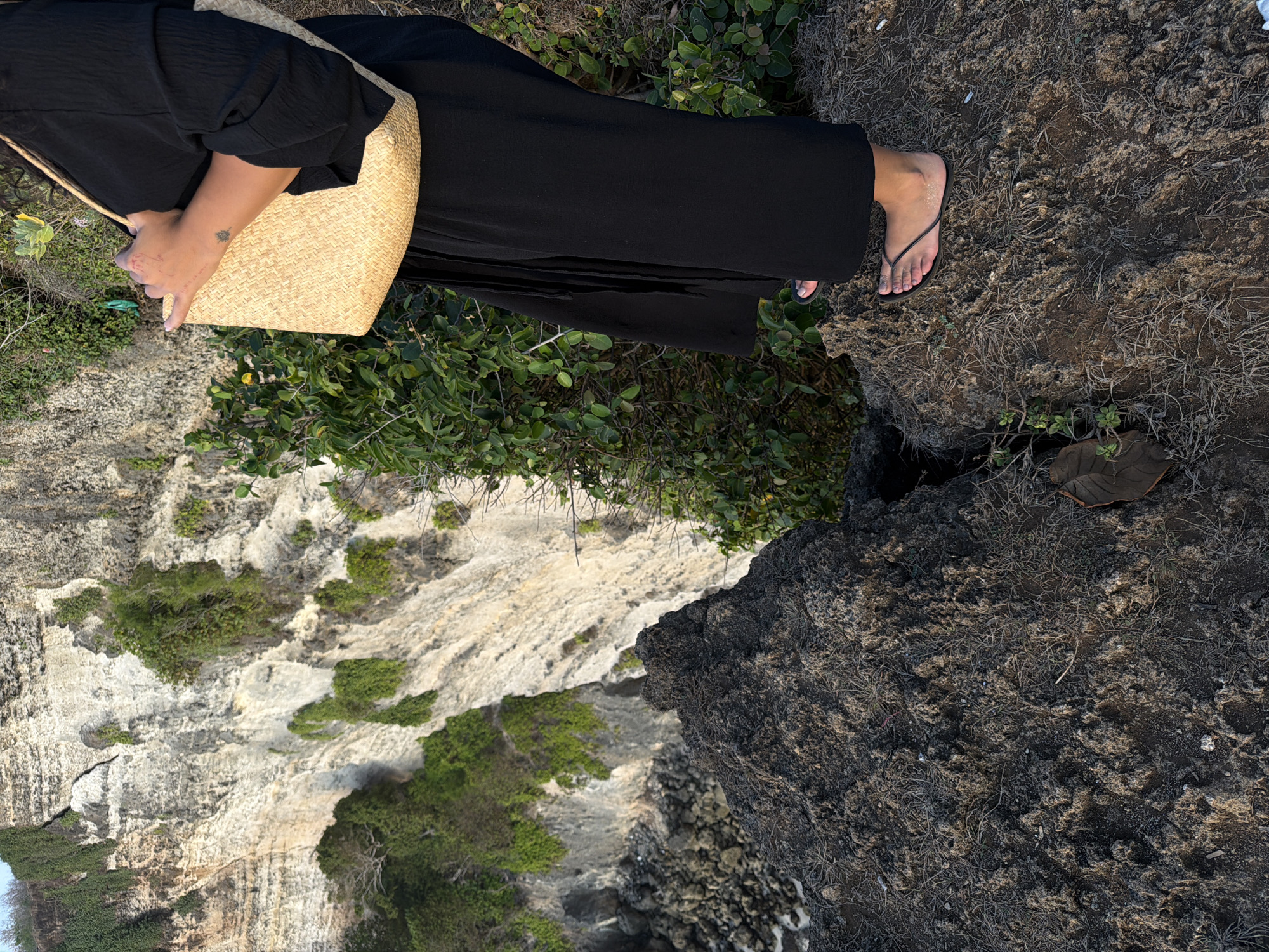 An individual stands on a rocky outcrop overlooking a steep cliff face in Kuta Selatan, Indonesia. The person is wearing a black dress and black flip-flops, and is carrying a woven straw bag. The cliff face is covered in green vegetation, and the ocean is visible in the distance. The lighting suggests it is either early morning or late afternoon.