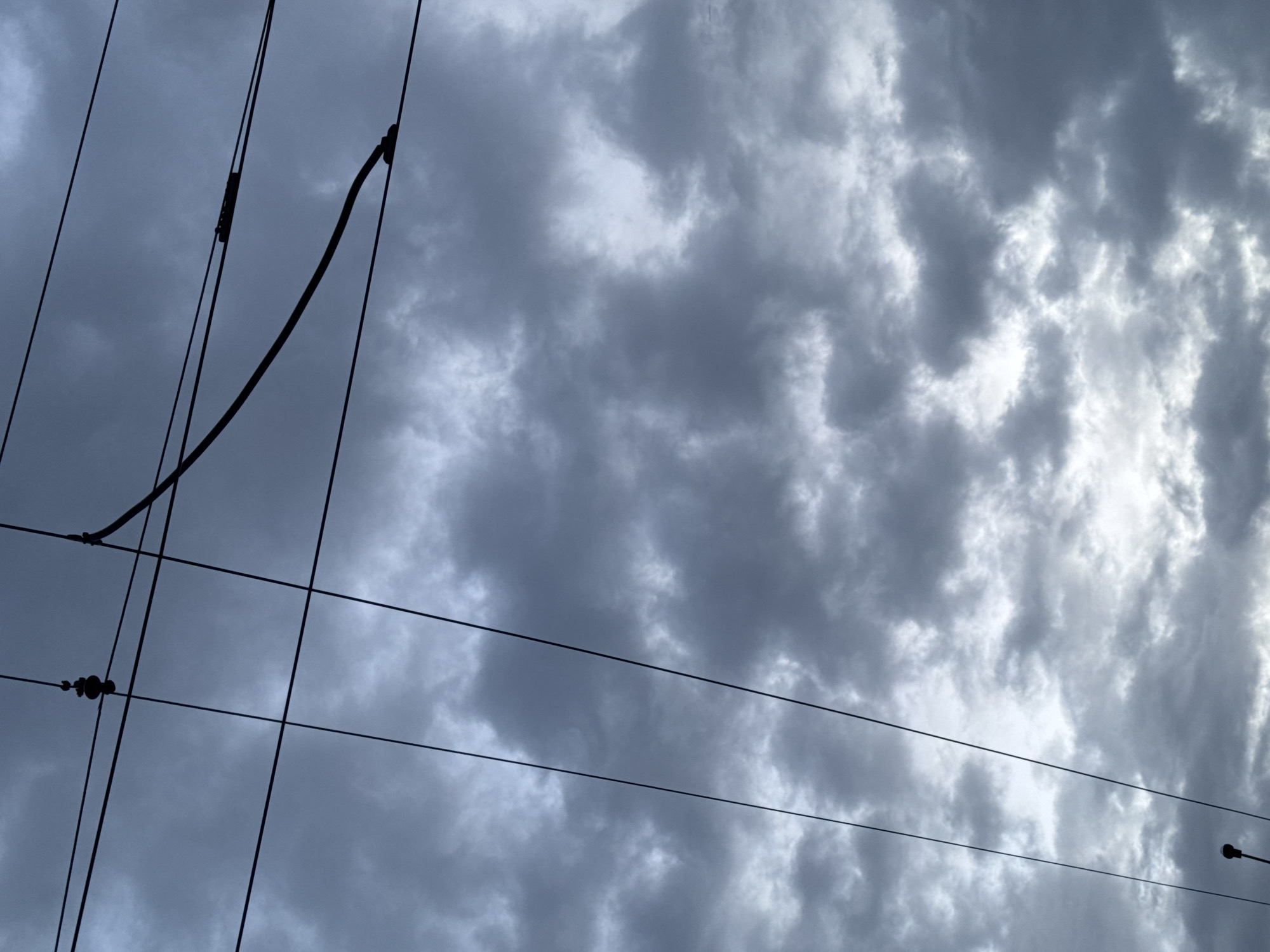 The image shows a cloudy sky with utility wires in the foreground. The clouds are gray and white, suggesting an overcast day. The utility wires are black and appear to be power lines or telephone wires. The location is likely Saint Petersburg, Russia, based on the typical urban landscape and architectural style often associated with the city.