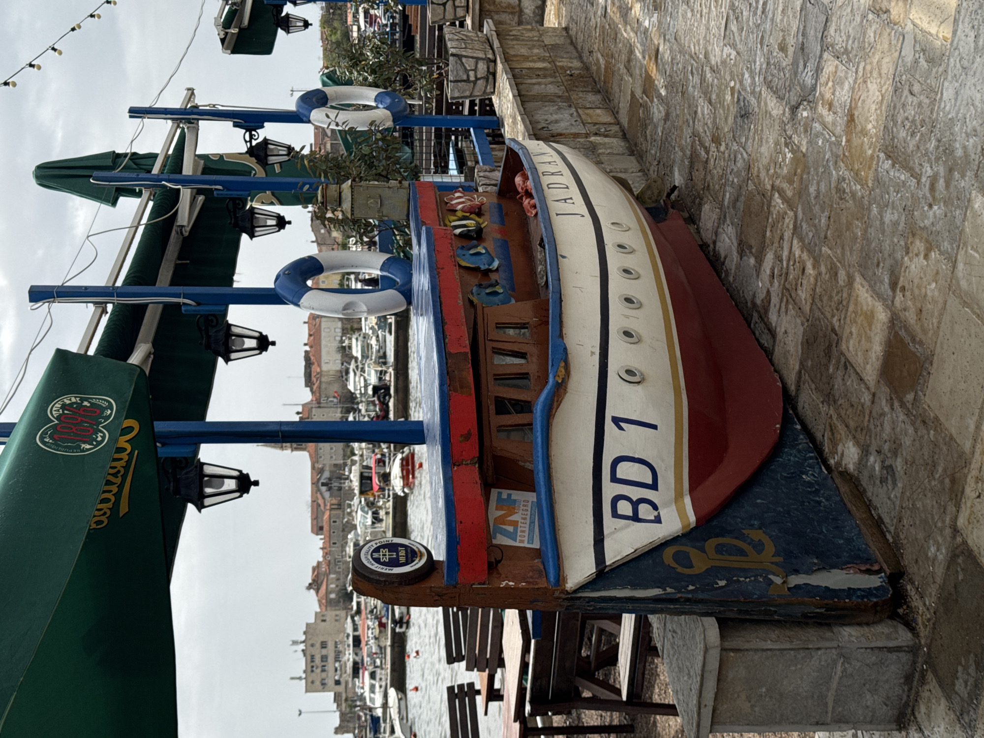 The image shows a weathered, decorative boat with the name "JADRAN" painted on its side, adorned with lifebuoys and positioned next to a stone wall. The boat has a red and white hull, with the letters "BD 1" painted on it. In the background, buildings and boats can be seen, suggesting a coastal setting. Above the boat, there is a green awning with the name "Nikšičko" printed on it, along with a blue structure that supports streetlights. The location is likely in Budva, Montenegro, given the boat's name and the background scenery. There are no people visible in the image.