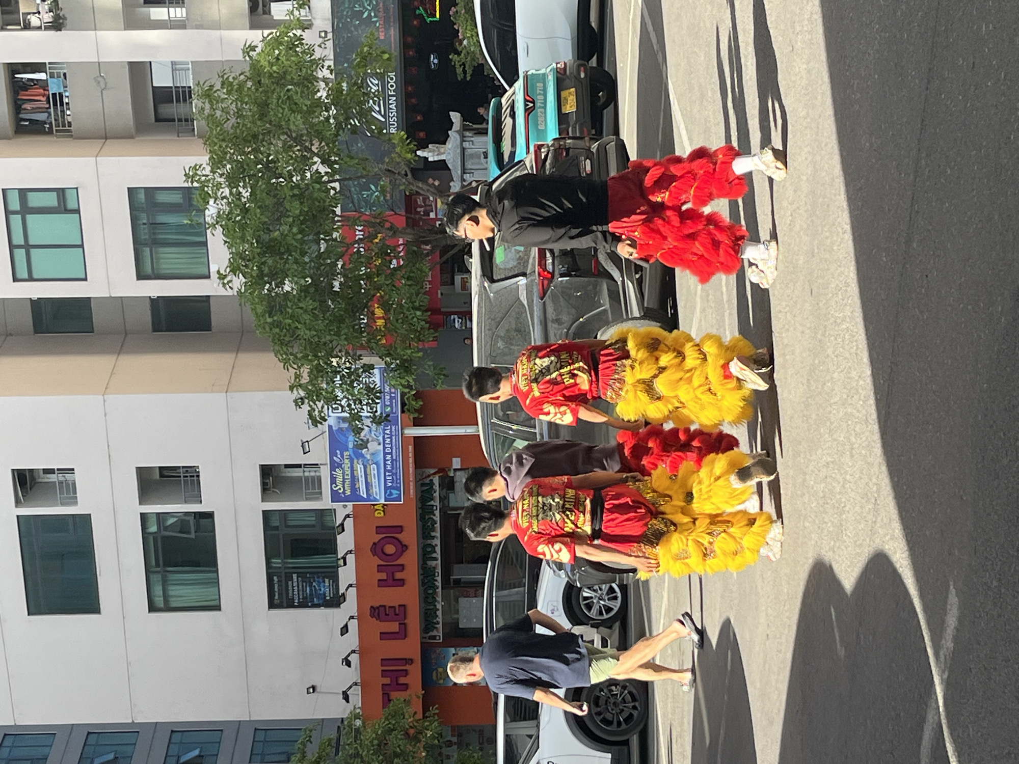 In a street scene in Bac Nha Trang Ward, Vietnam, a procession of performers in traditional red and yellow lion dance costumes moves along the sidewalk. Behind them, people, including a Caucasian man in a dark shirt and shorts, watch and follow. Vehicles line the street, some partially obscured by the performers. A building with windows, balconies, and an advertisement for Viet Han Dental is visible in the background. A sign reads "THỊ LỄ HỘI WELCOME TO FESTIVAL." The image is brightly lit, with shadows cast on the pavement.