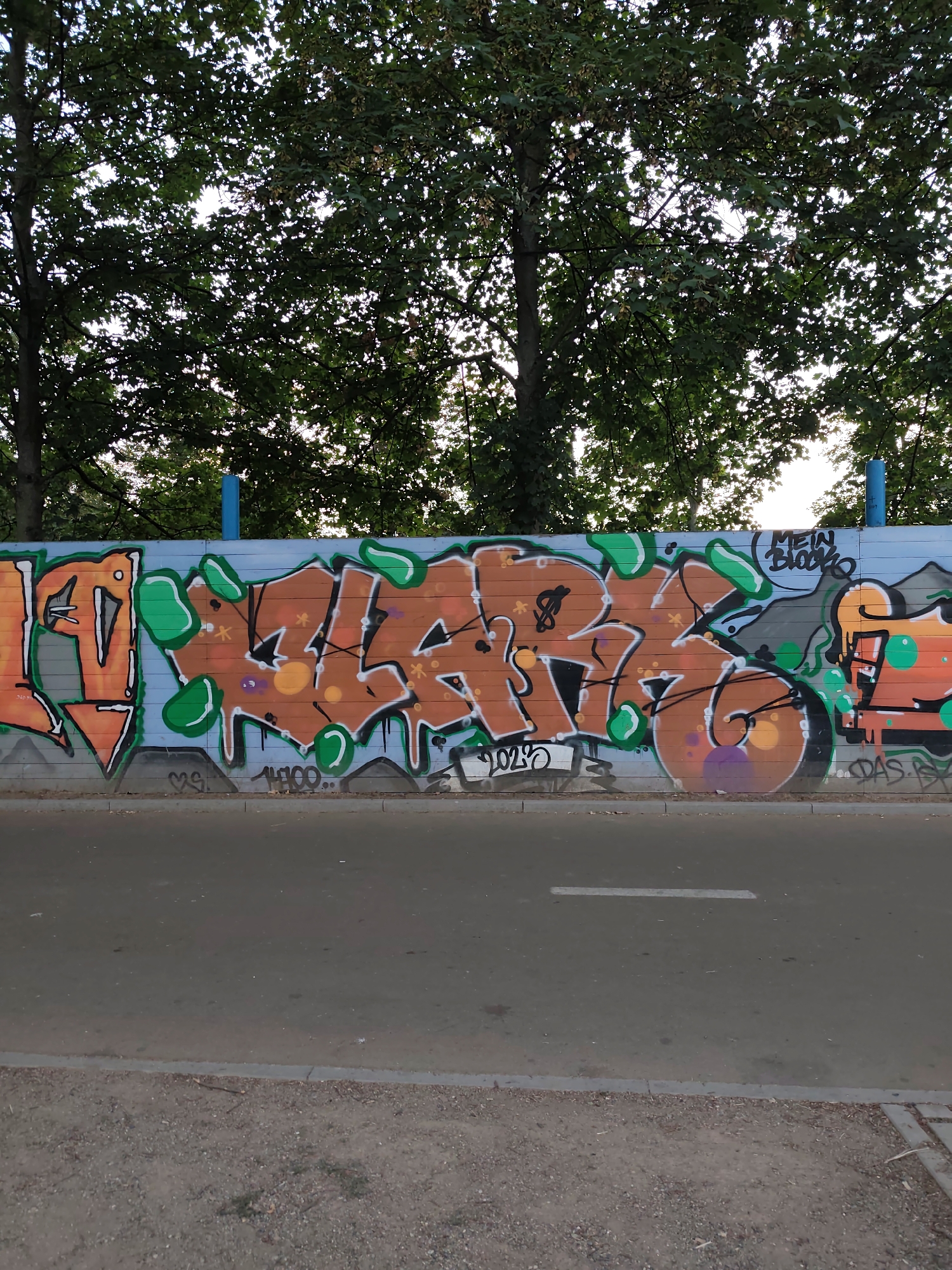 The image shows a graffiti-covered wall in Prague, Czechia. The wall is made of horizontal wooden planks painted blue, with the graffiti predominantly in orange and green. The graffiti features large, stylized letters and abstract shapes, with some smaller tags and details. A road runs along the base of the wall, with a sidewalk and some gravelly ground visible in the foreground. Trees with green foliage are visible above the wall, suggesting an outdoor setting. There are no people or animals in the image. The scene appears to be a public space, possibly a park or a street with a designated graffiti area.