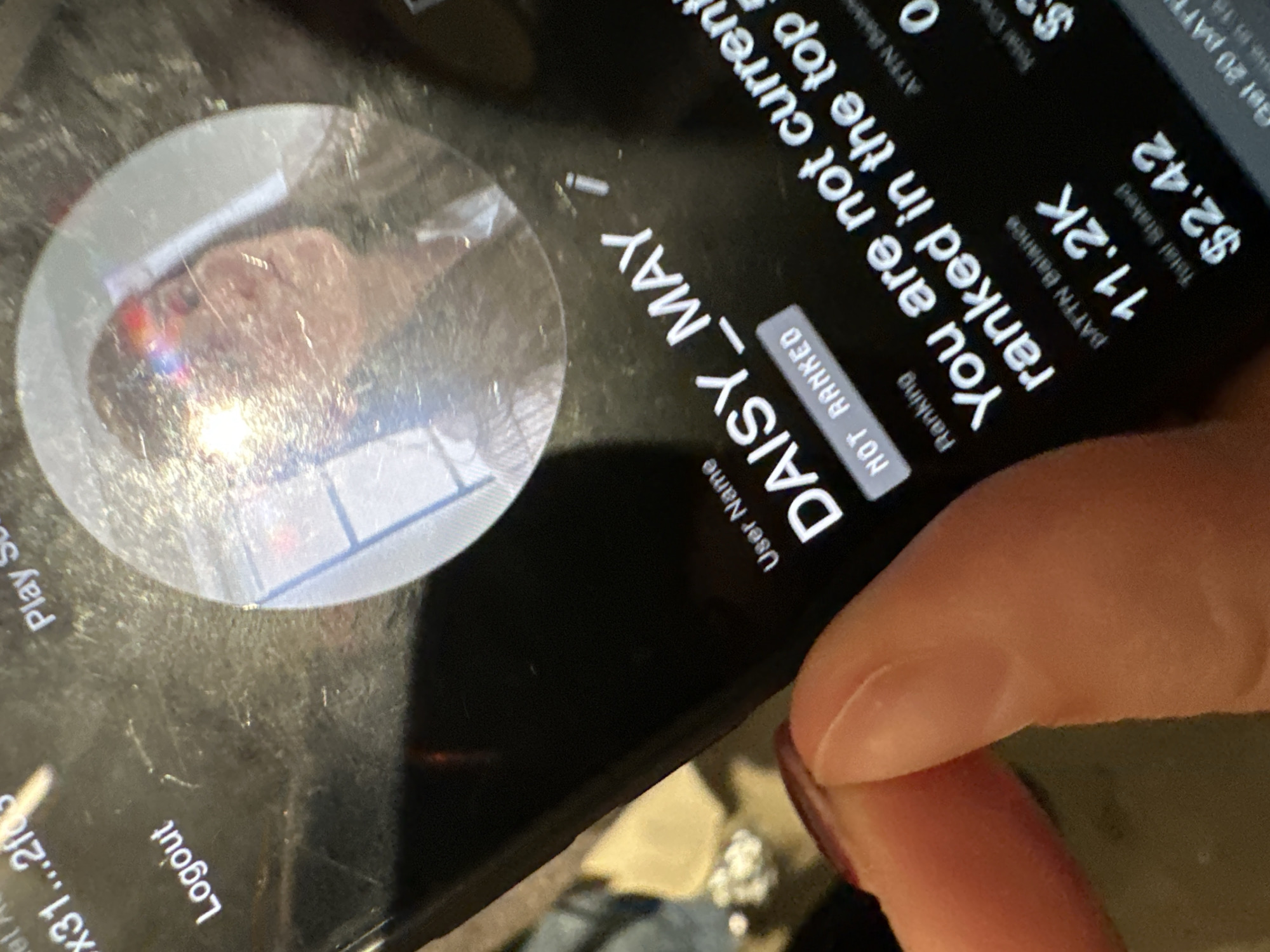 The media file captures a close-up, angled view of a smartphone screen being held by a person. A finger is visible in the bottom right corner, suggesting interaction with the device. The screen displays an application interface with a dark background and light-colored text. A circular profile picture area on the left shows a blurry reflection of a person, possibly the photographer, with light glare over the face. The screen itself appears to have smudges or scratches.
The app shows user account information. Visible text includes a "User Name" identified as "DAISY_MAY". Under "Ranking", it states "NOT RANKED". A partial message reads, "You are not current ranked in the top". Financial or balance information is displayed, with "ATTN Balance 11.2K" and "Total Staked $2.42". Other text snippets visible are "Logout" and "Play S" on the left, and partially "Get 20 BATTN" and "$3" on the top right. The activity depicts someone viewing their profile or dashboard within an application, likely a gaming or social platform, given the ranking and balance details. The setting appears to be indoors, with no specific location cues within the image, though the provided context suggests London, United Kingdom.