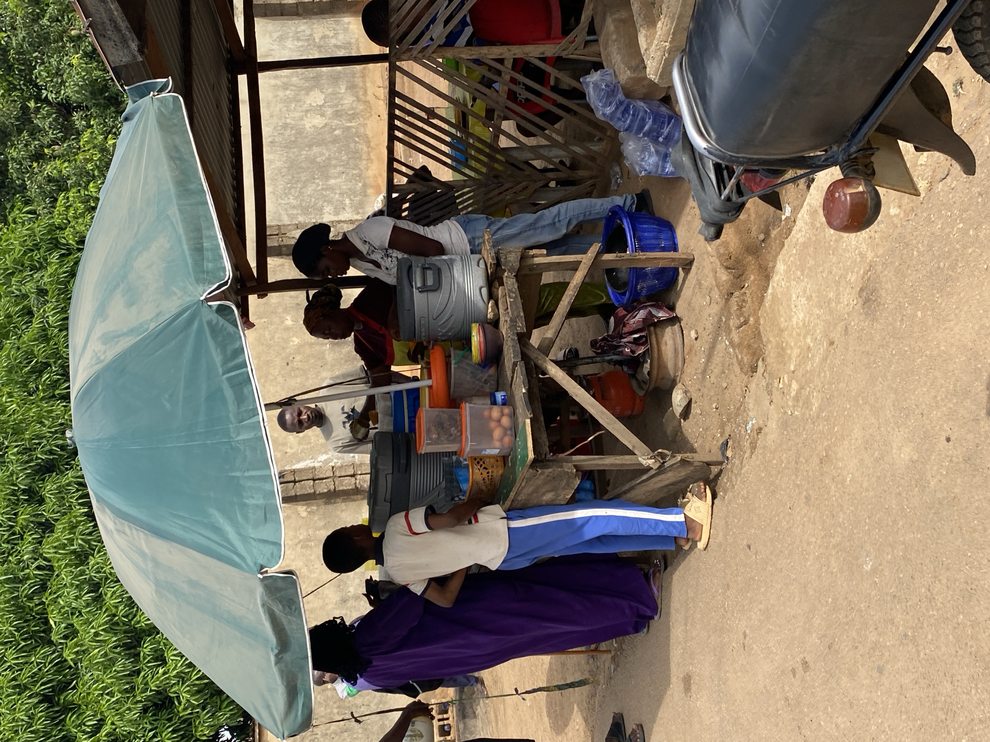 The image shows an outdoor scene in Narayi, Nigeria. Under a makeshift shelter with a large blue umbrella, people are gathered around a food or refreshment stand. A couple of women seem to be working at the stand, possibly serving customers. A motorcycle is parked nearby. There are various items on the stand, including bottles, containers, and a cooler, suggesting a variety of offerings. The environment is typical of a local marketplace or roadside vending area.